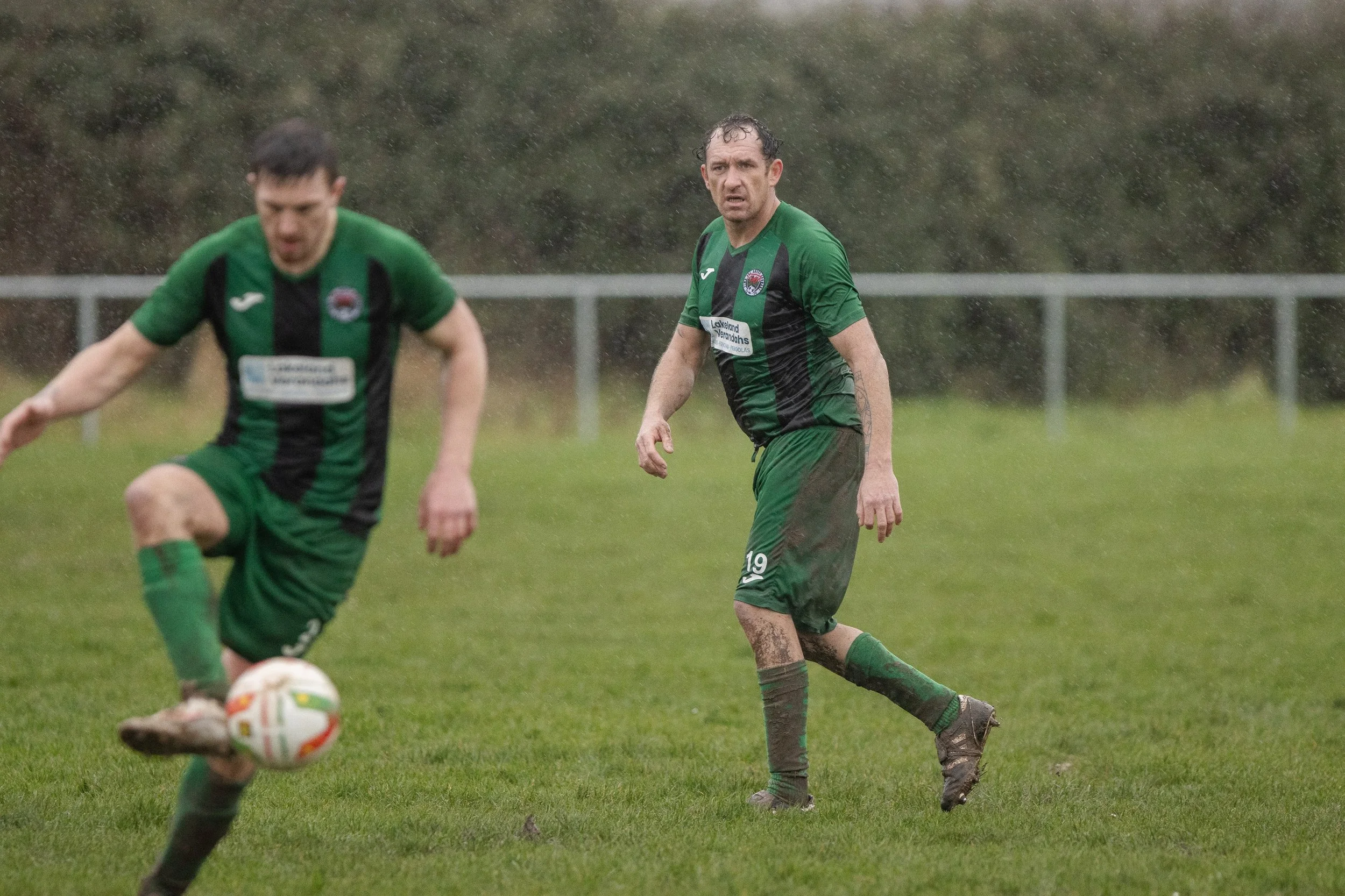 Two soccer players in green and black uniforms playing on a rain-soaked field, one about to kick the ball, and the other looking towards the camera with muddy clothes.