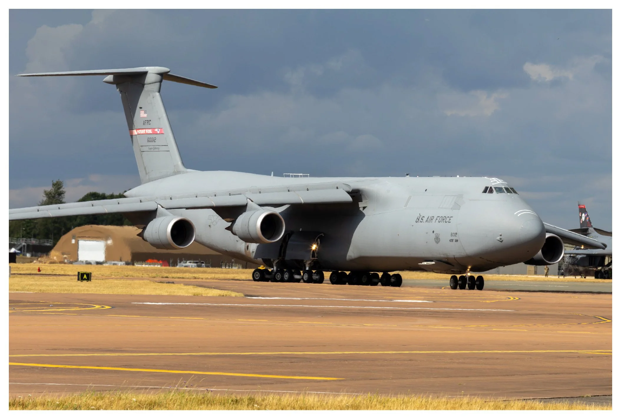 Large gray military cargo airplane on the runway at an airport, with the sky and some trees in the background.