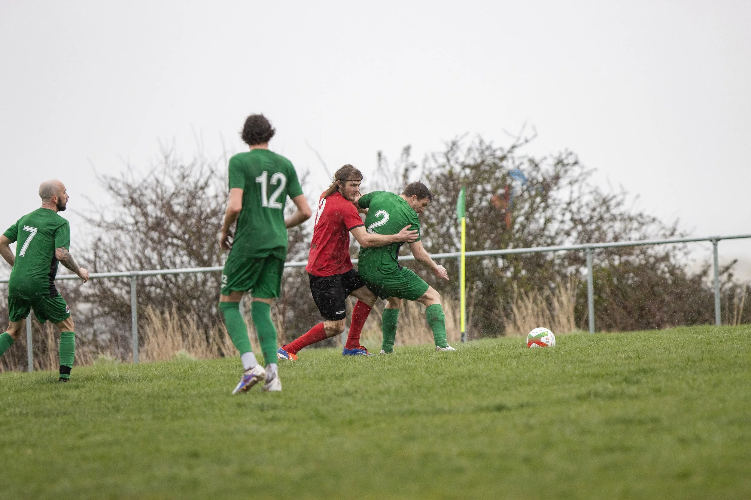 A soccer match in rain, players in green and red uniforms fighting for the ball near the corner flag.