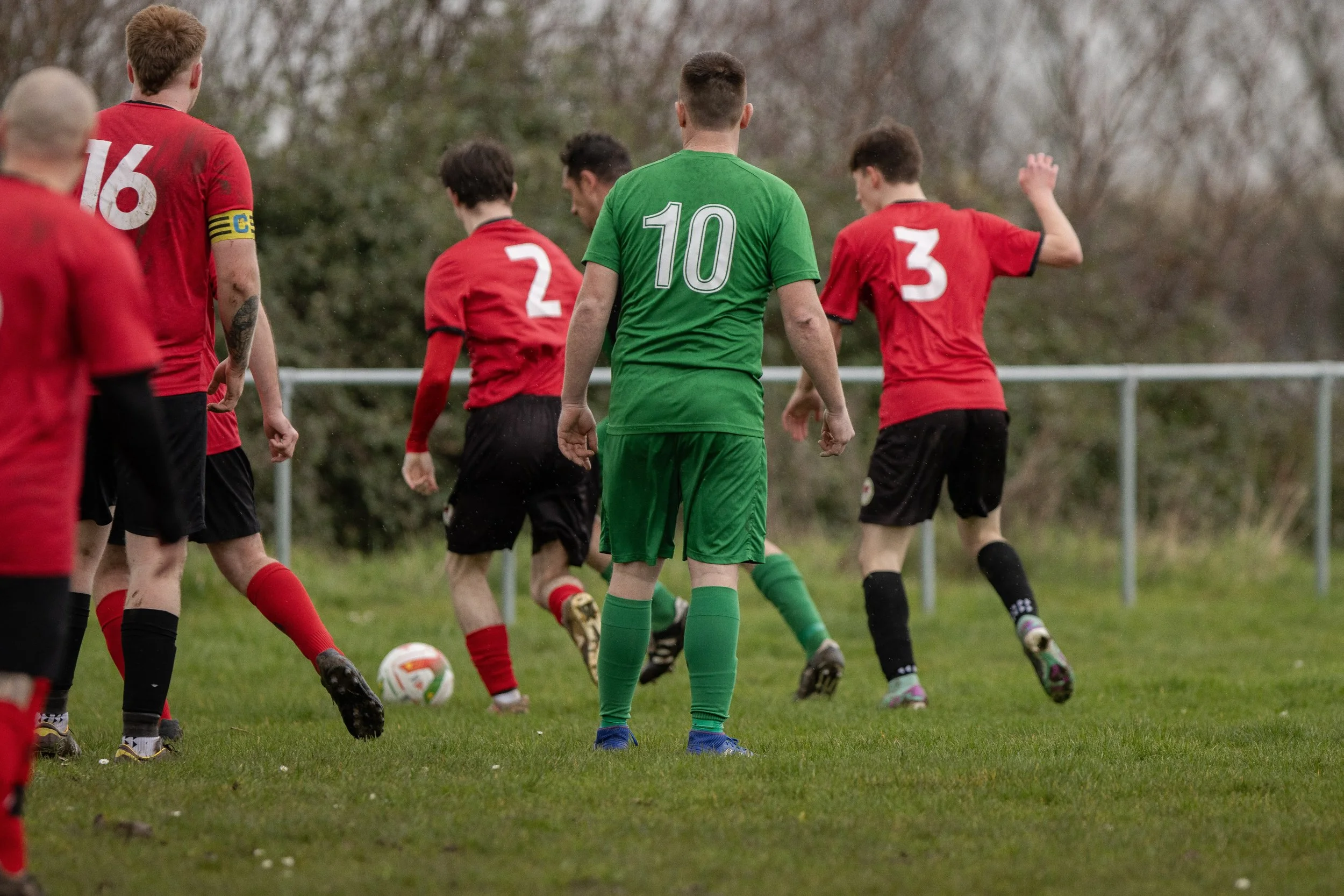 Soccer players in red and green uniforms compete on a grassy field, with one player in a green jersey and shorts facing away from the camera and others in red jerseys and black shorts surrounding a soccer ball.