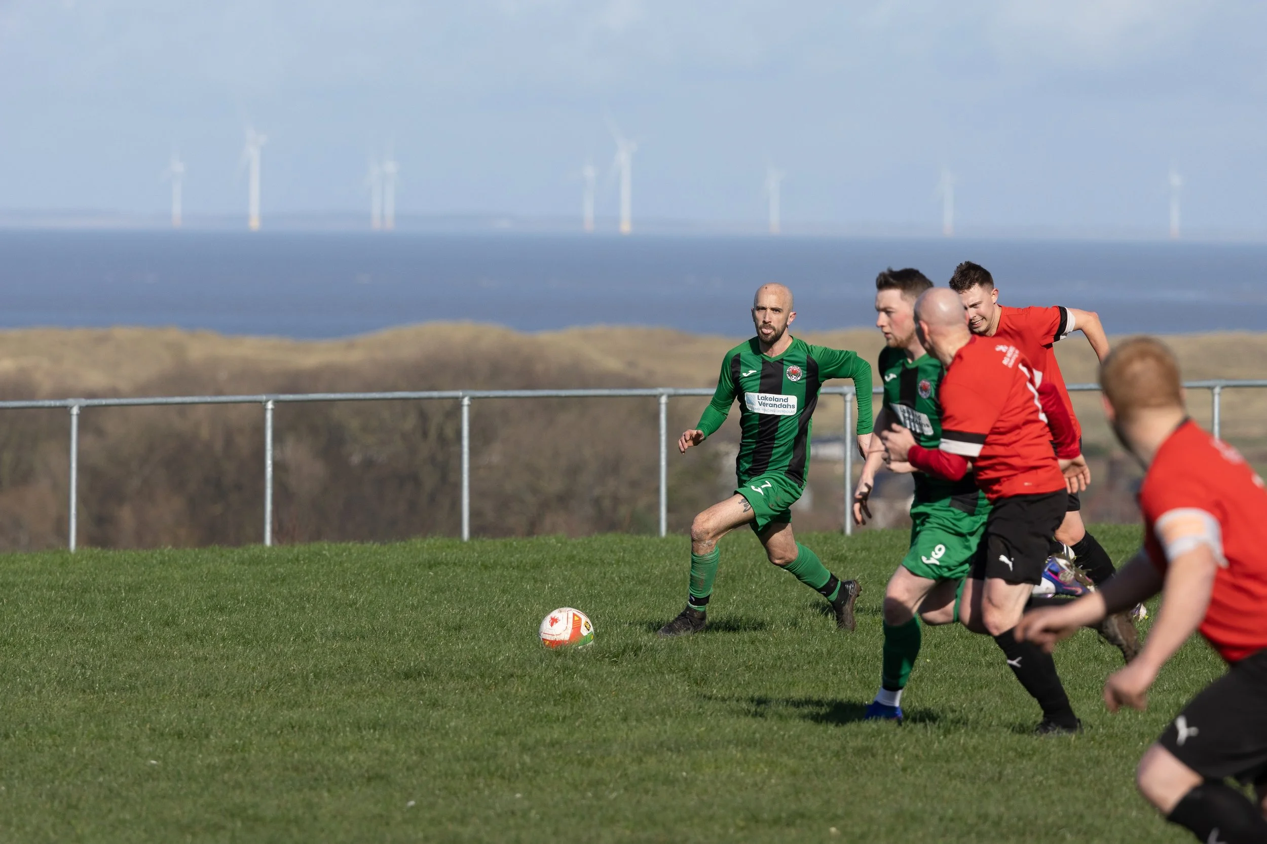 Soccer game with players in green and red jerseys on a grassy field near a body of water, with wind turbines in the background under a partly cloudy sky.