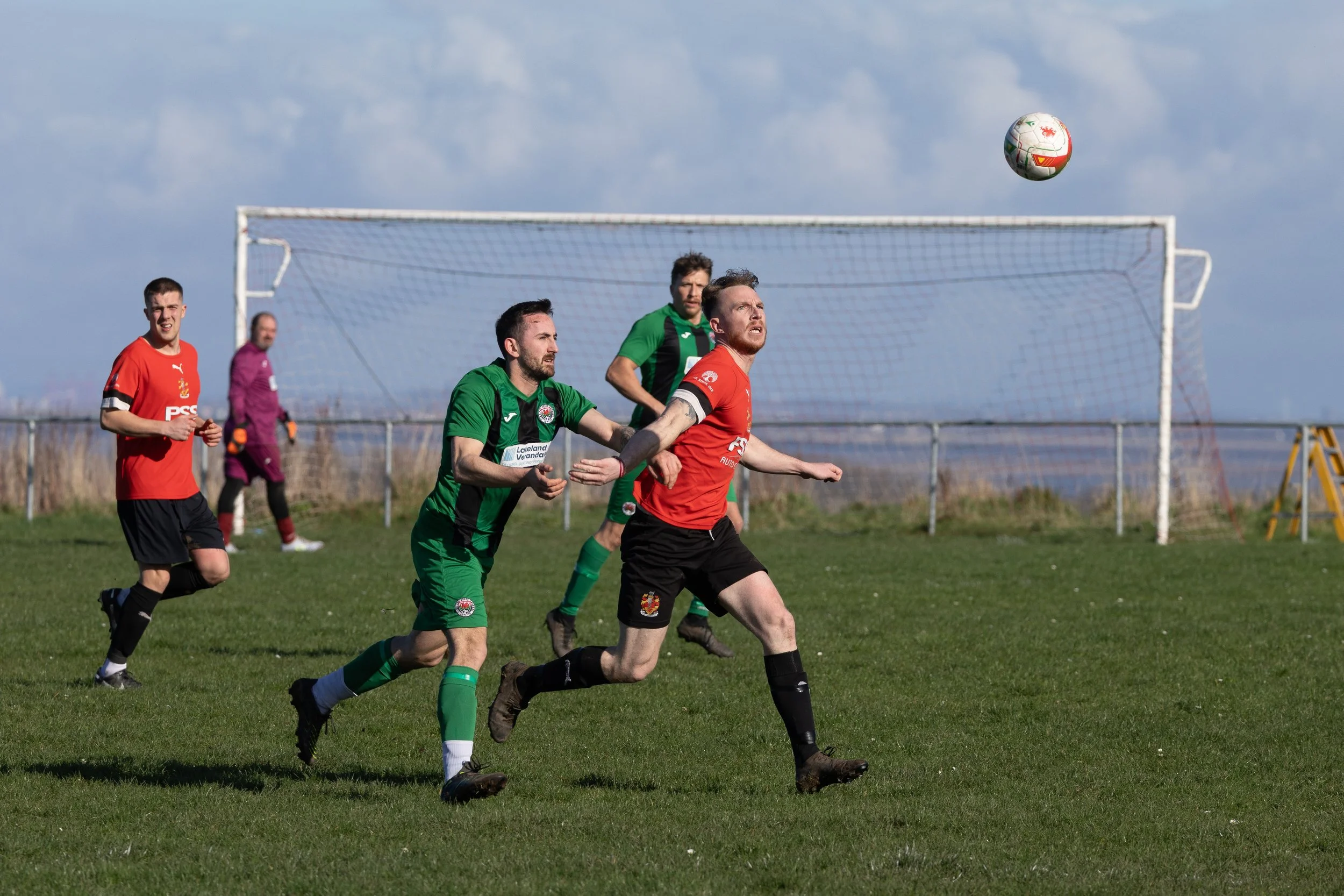 Soccer players in action on the field, with two players chasing the ball and a goalkeeper in the background, under a partly cloudy sky.