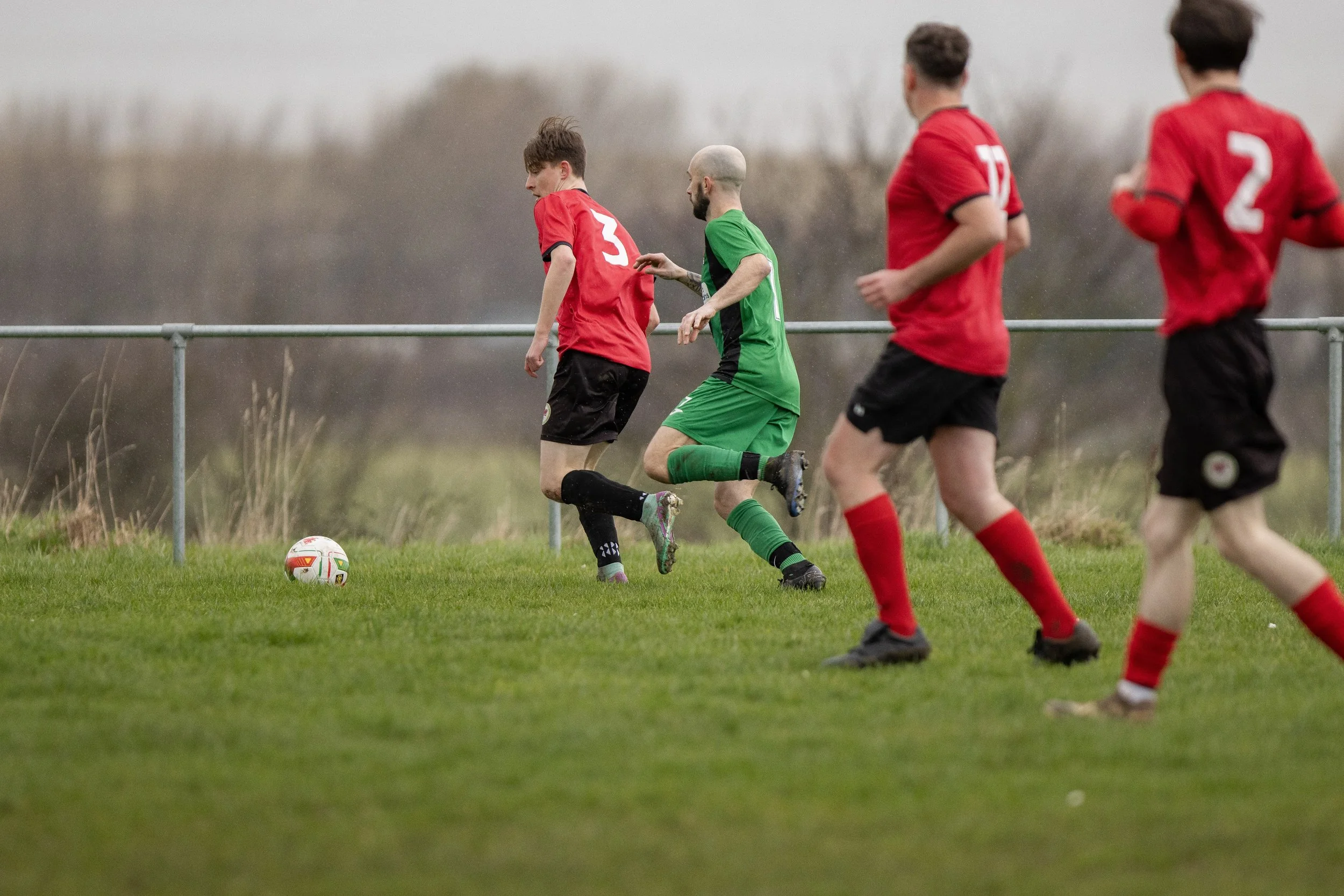 Soccer game with four players on a grassy field, one in a green jersey and three in red jerseys, racing towards the ball.