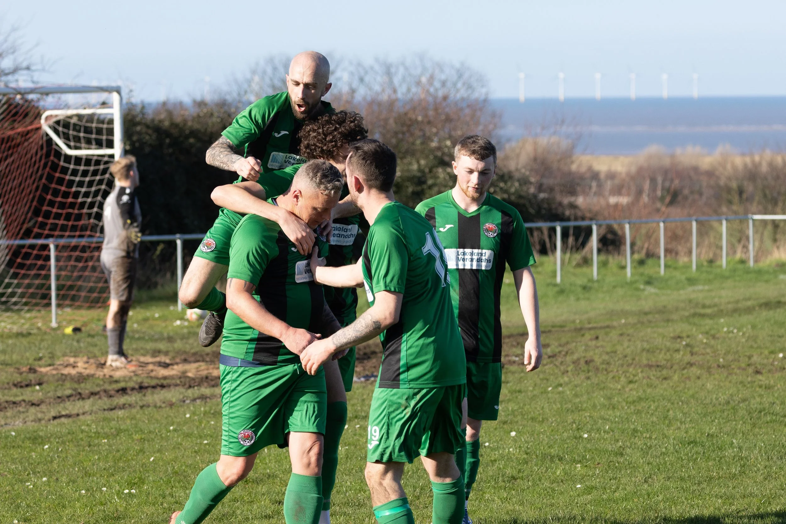 Soccer players in green and black uniforms celebrating on a grassy field, with goalpost and fence in the background.