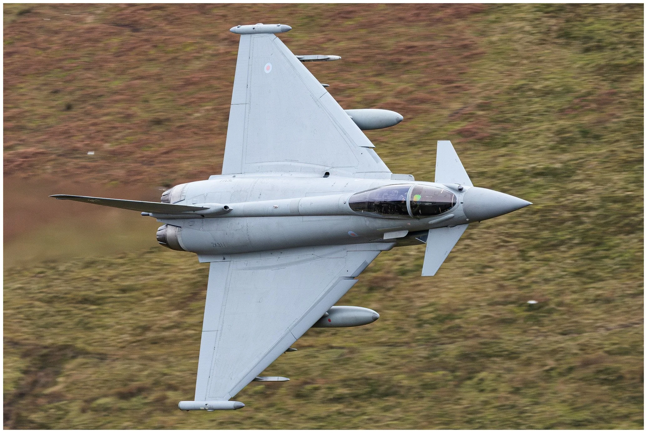 A military fighter jet flying low over grassland, viewed from above.