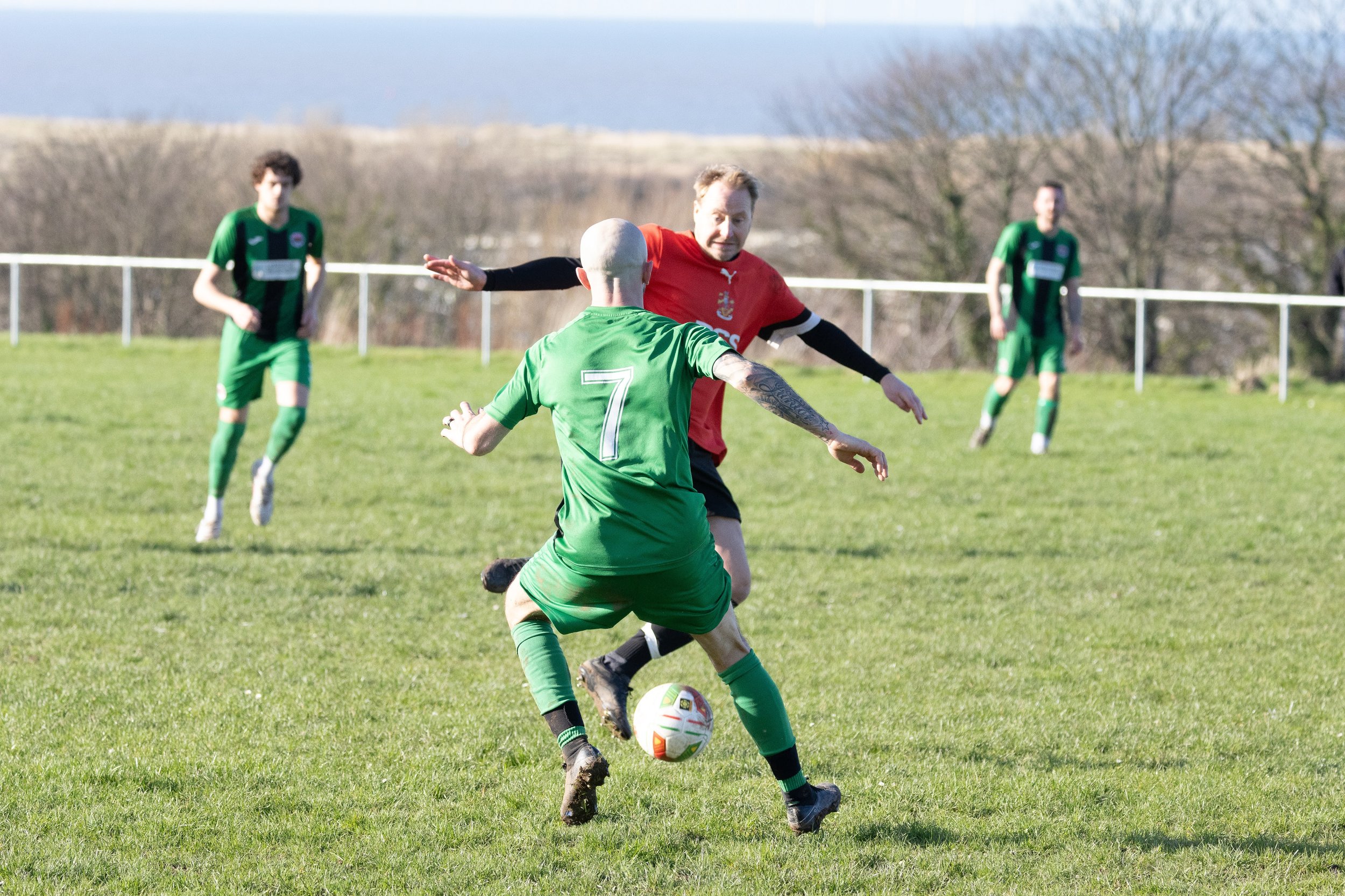 Two soccer players, one in a green uniform with the number 7, and the other in a red uniform, are competing for the ball on a grassy field. Two more players in green uniforms are in the background, with a fence, trees, and a body of water behind them