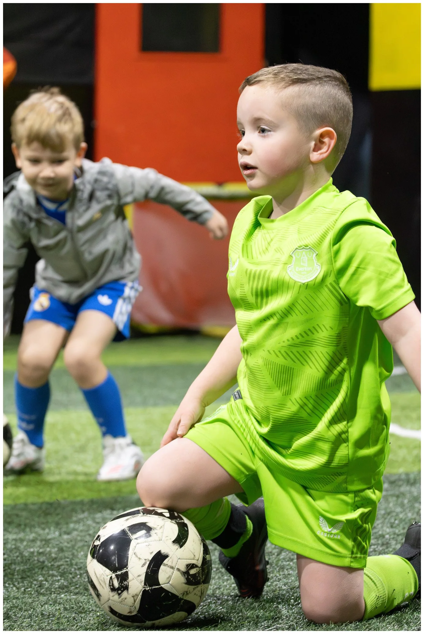 A young boy in a bright green soccer uniform kneels on an indoor soccer field with a soccer ball at his knee, while another child in gray and blue sportswear is blurred in the background.