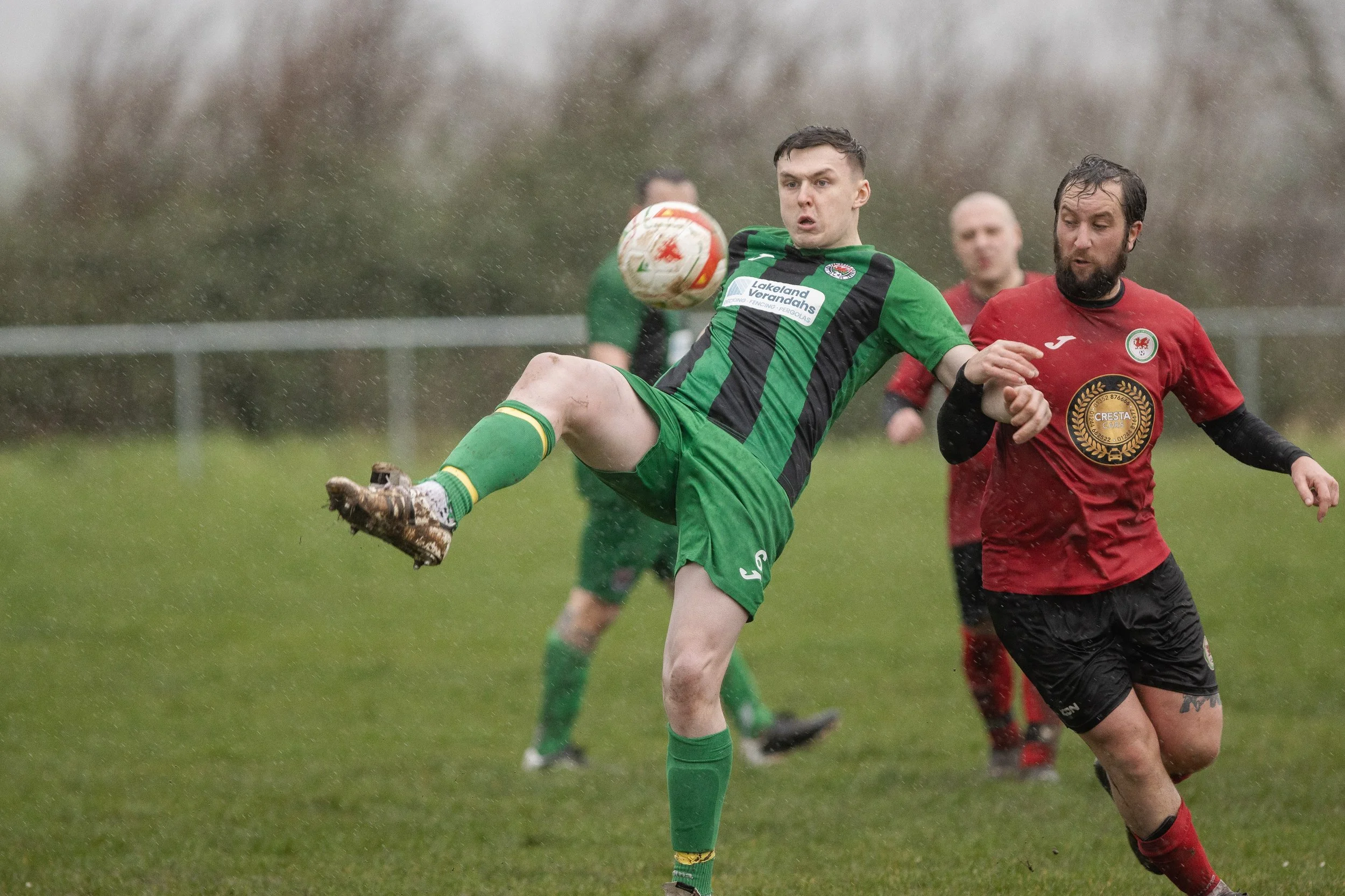 A soccer player in a green and black uniform is kicking a ball during a match, with an opponent in a red and black uniform nearby. Rain is falling on the field.