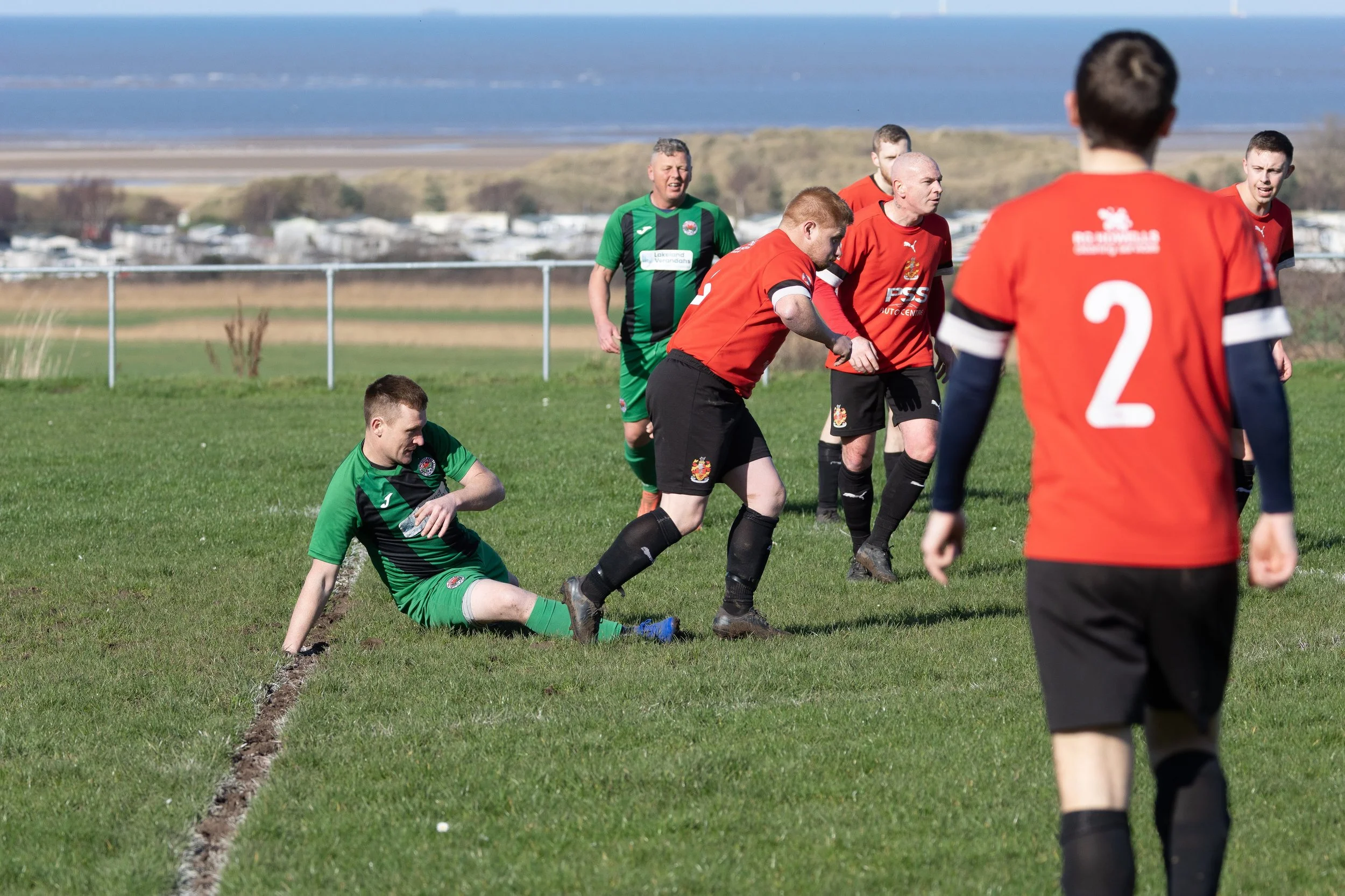 Soccer players on a field, with some in red jerseys and one in green, during a match near a coastline with water and cars in the background.