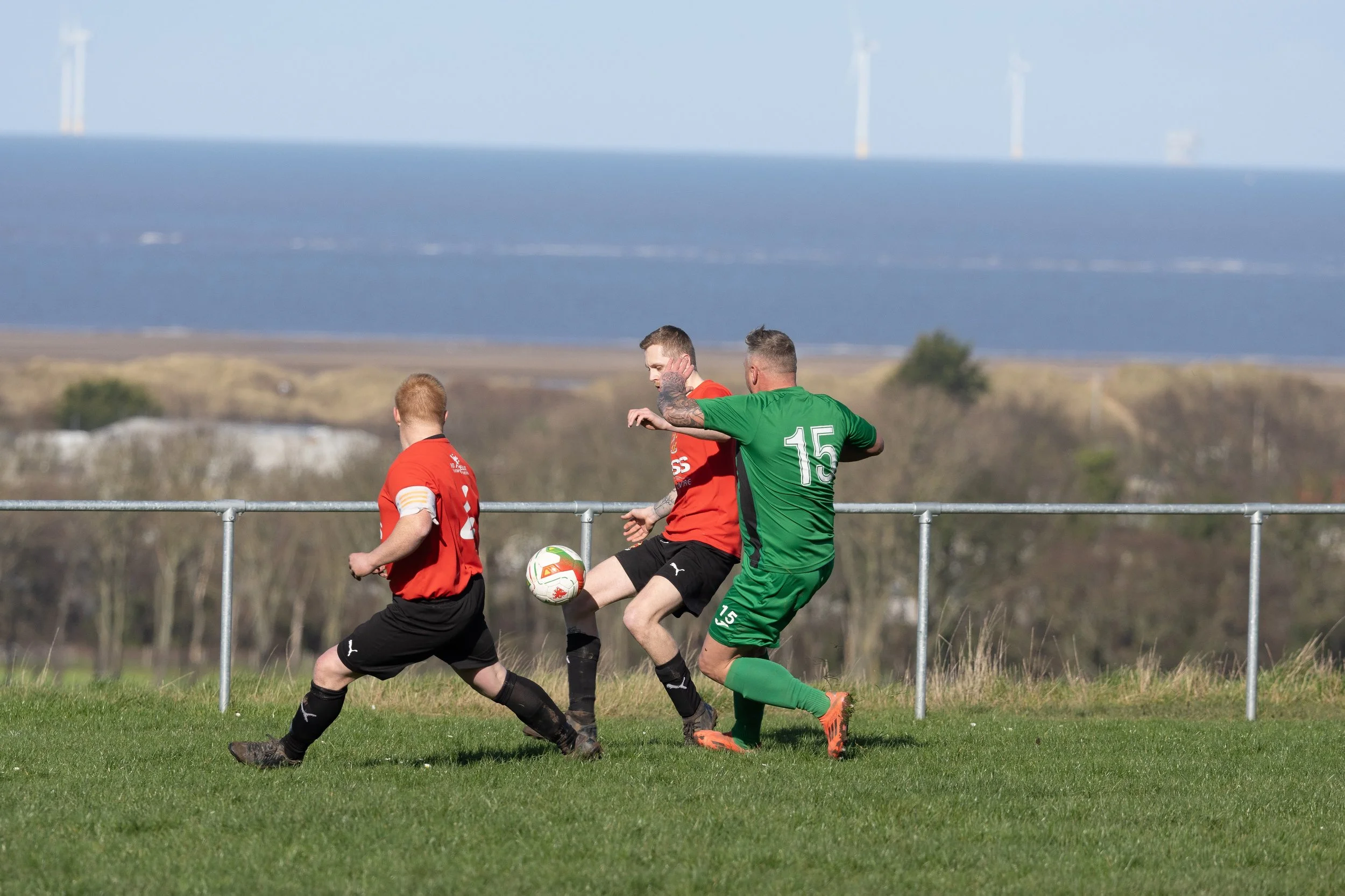Three soccer players in red and green jerseys playing soccer on a grassy field, with a seaside landscape and wind turbines in the background.