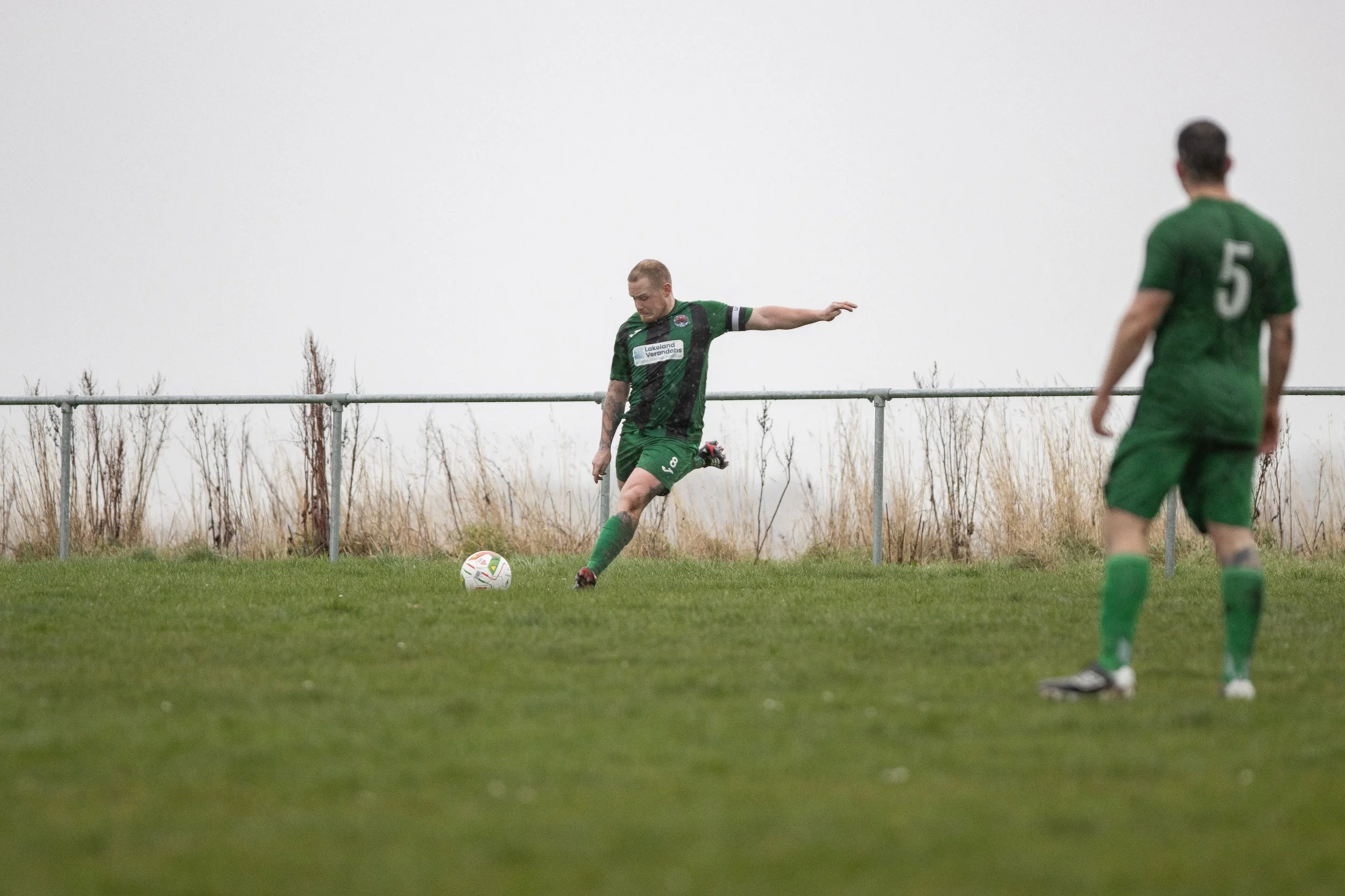 Two soccer players in green uniforms on a grassy field, one kicking a soccer ball while the other observes.