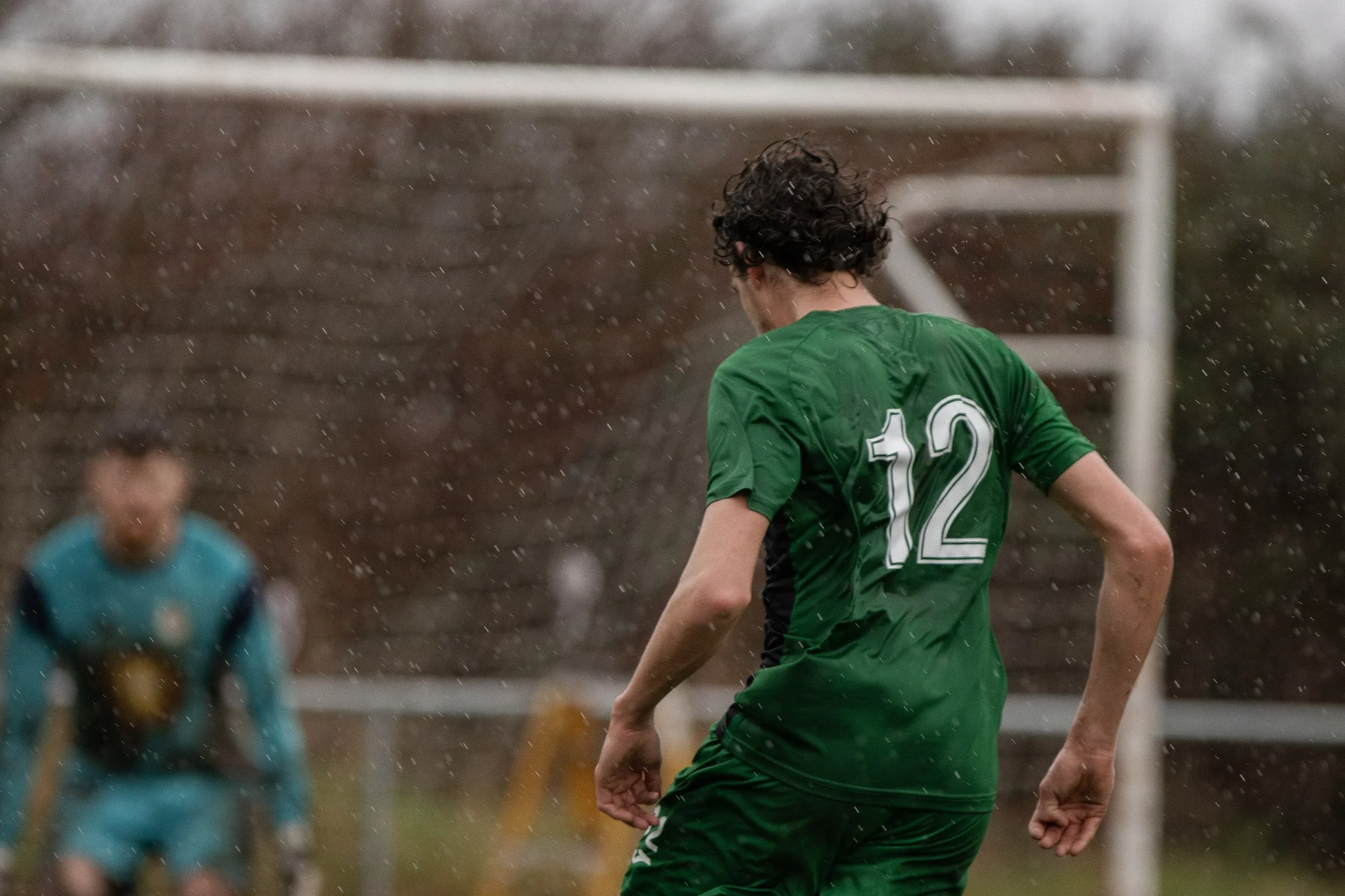 A soccer player in a green jersey with the number 12 is on a field during rain, with a goalkeeper in the background.