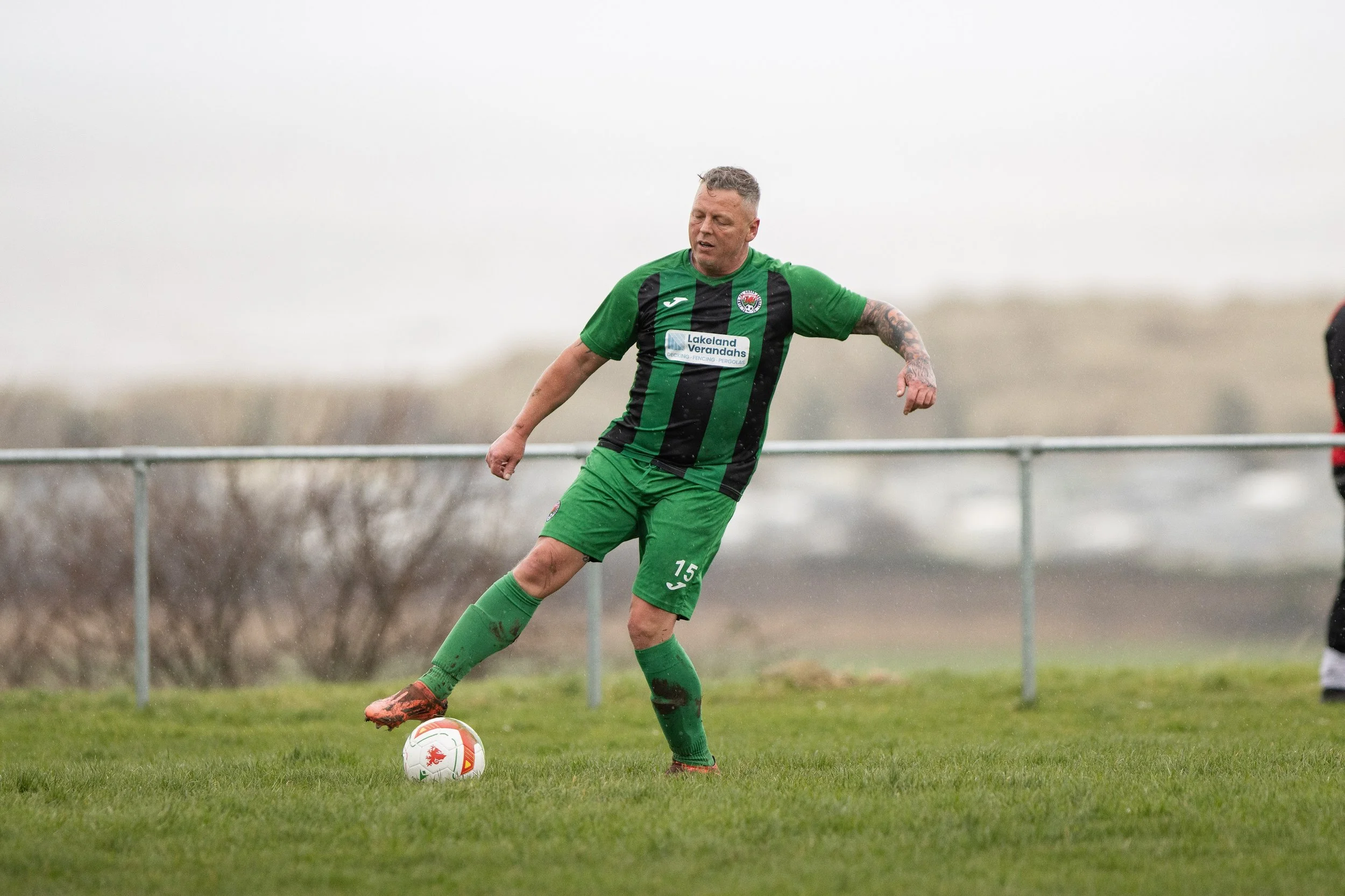 A man playing soccer on a grassy field, wearing a green and black uniform, kicking a soccer ball. He has short, gray hair and tattoos on his left arm.