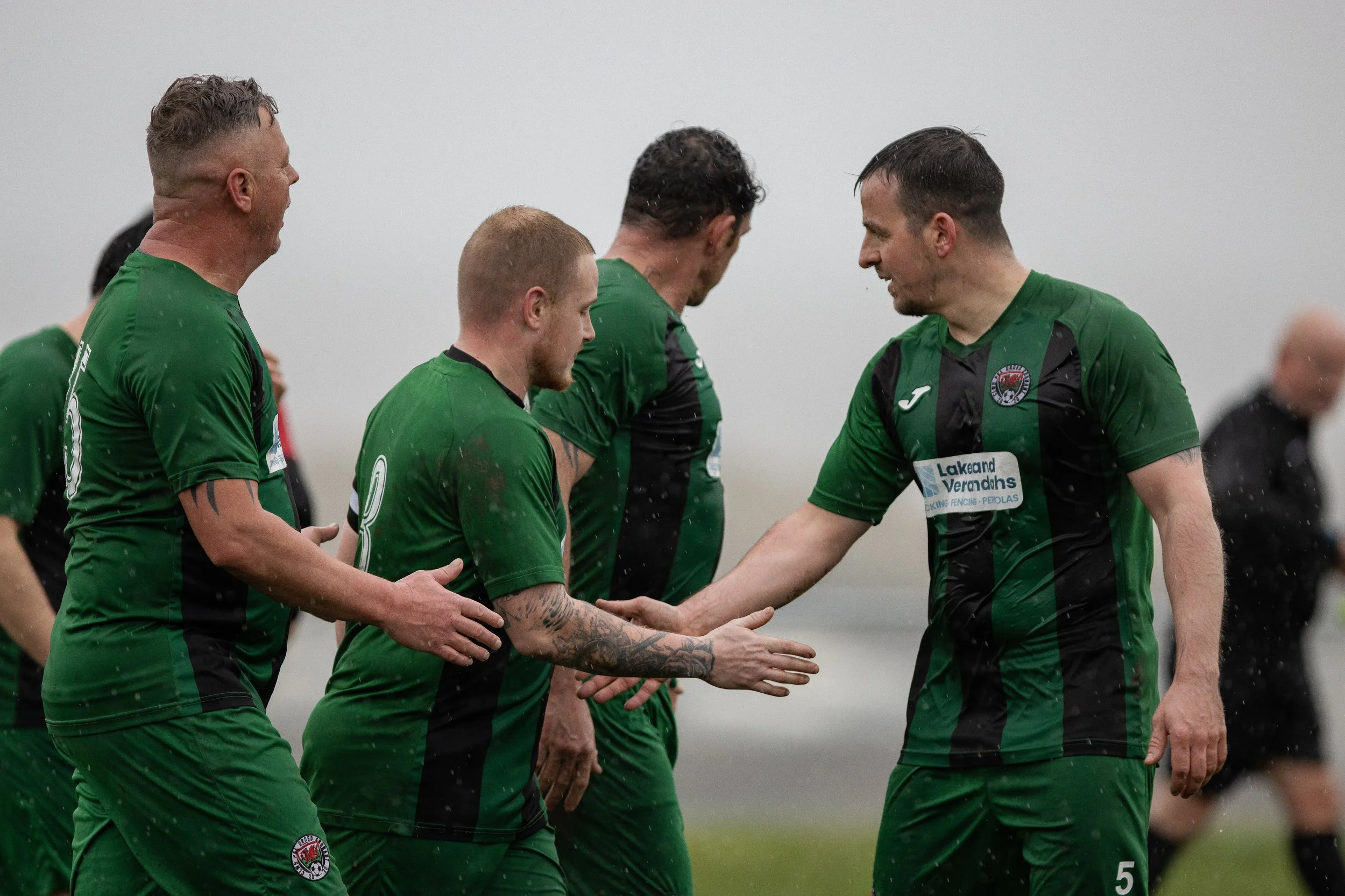 Soccer players in green jerseys shaking hands before a match on a rainy day.
