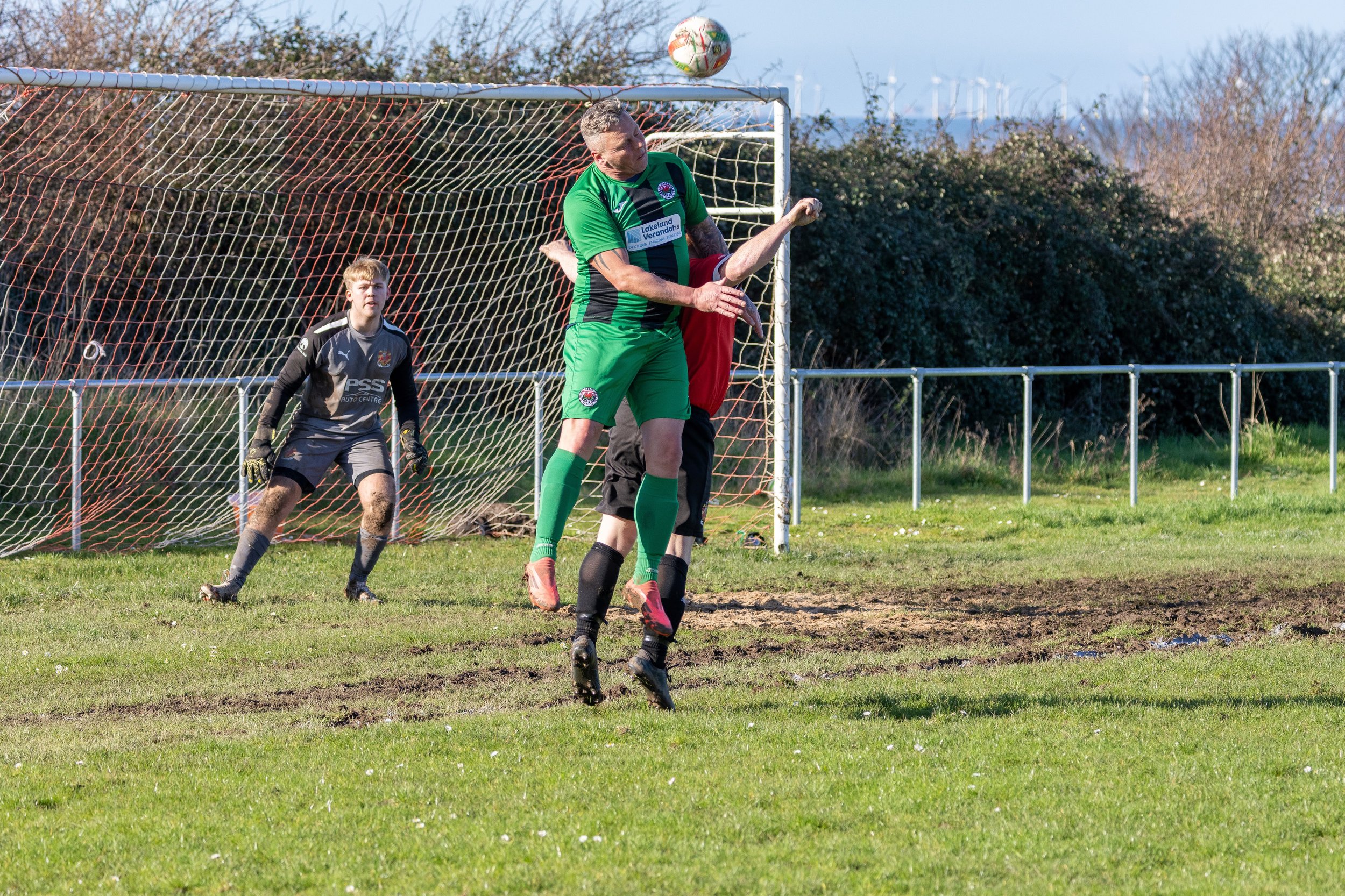 Soccer player in green jersey jumping to head the ball near the goal with goalkeeper and another player behind. Goalpost, net, grassy field, and bushes in background.