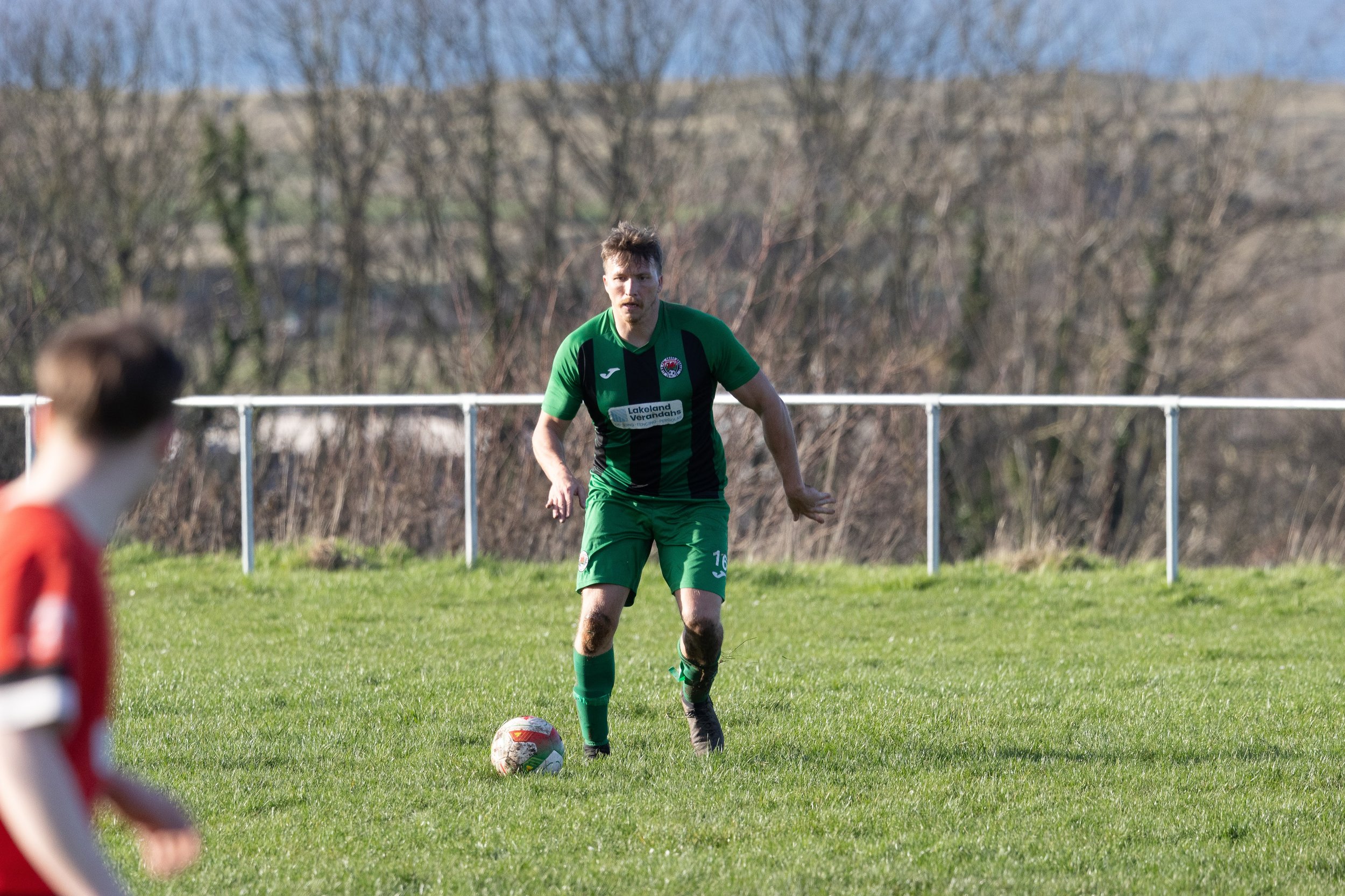 A soccer player in a green uniform preparing to kick a soccer ball on a grassy field, with another player in red in the foreground.