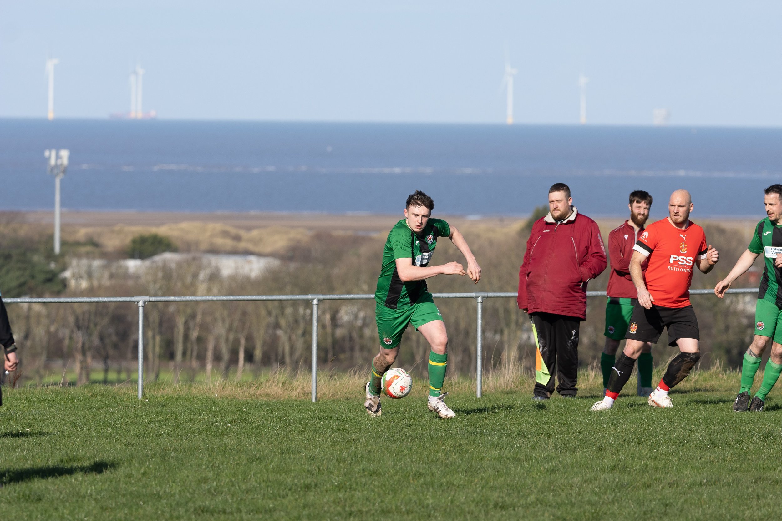 A soccer player in green uniform running with the ball during a game, with several other players and a coach on the sidelines, coastal landscape with wind turbines in the background.