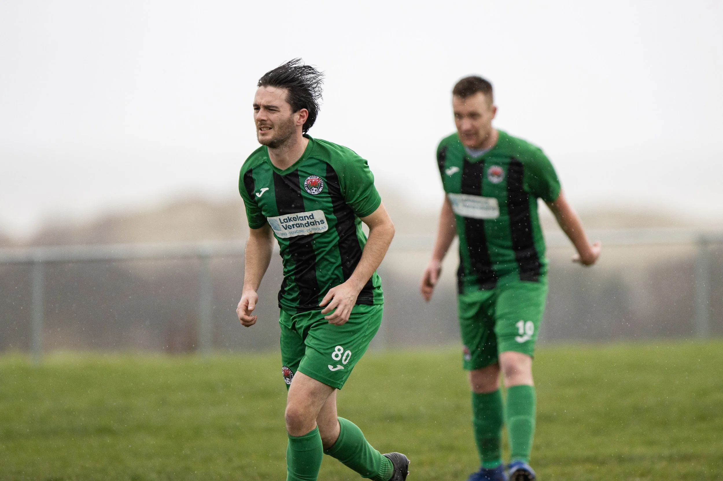 Two soccer players in green and black jerseys on a rainy field during a game, with one player in the foreground grimacing and another in the background.
