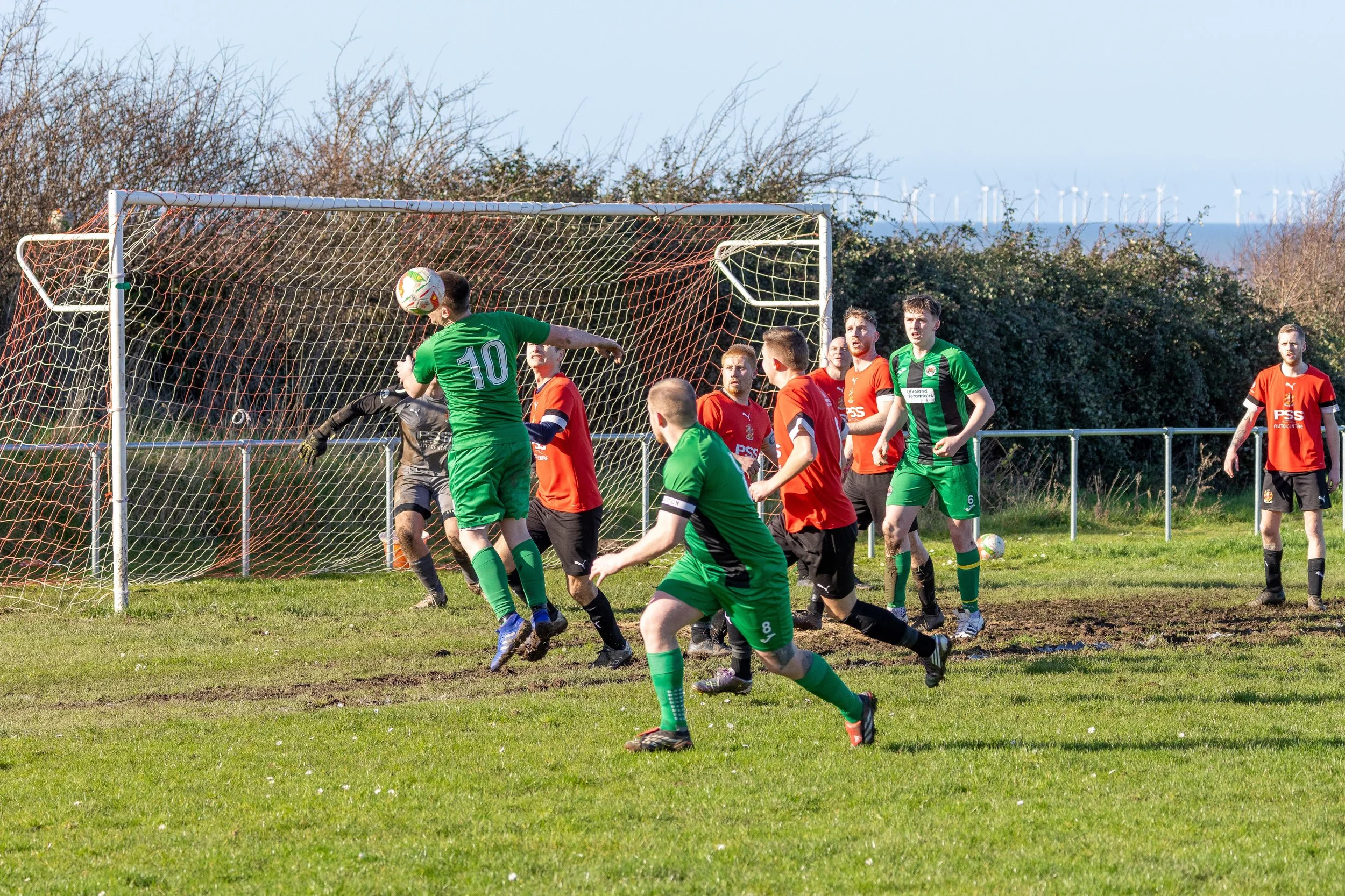 Soccer players in green and red jerseys compete for a ball near the goal on a grassy field.