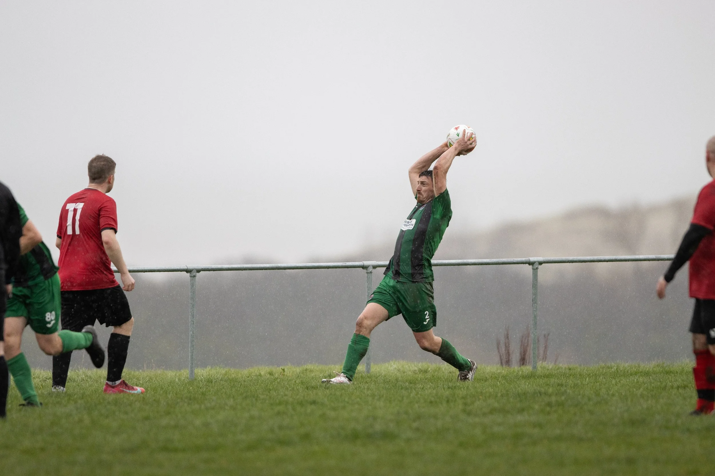 A soccer goalkeeper in green uniform catching a ball with both hands during a game in rainy weather, with players in red and green uniforms nearby on a grassy field.