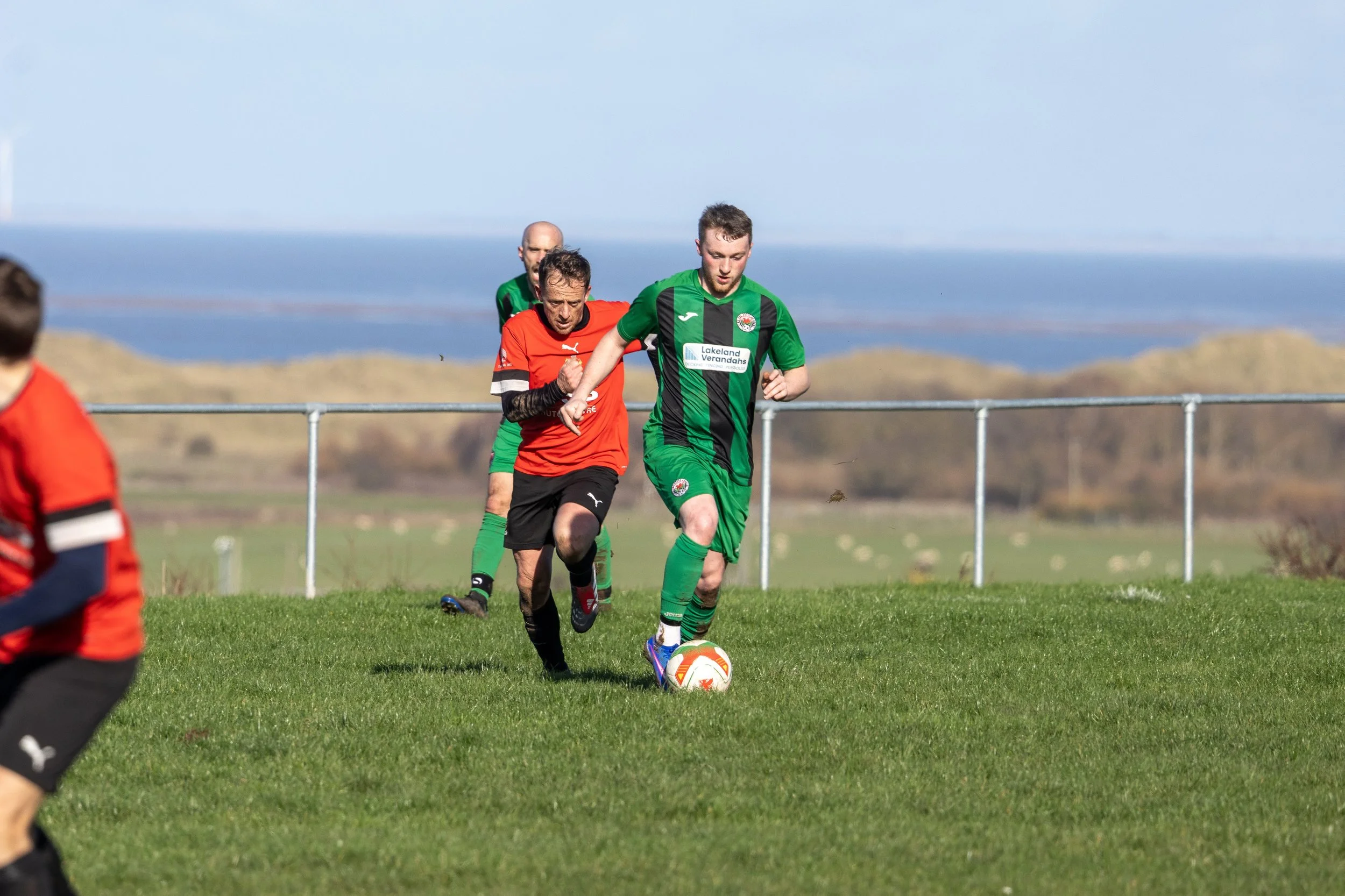 Soccer players in green and red uniforms chase the ball on a grassy field with distant hills and a body of water in the background.