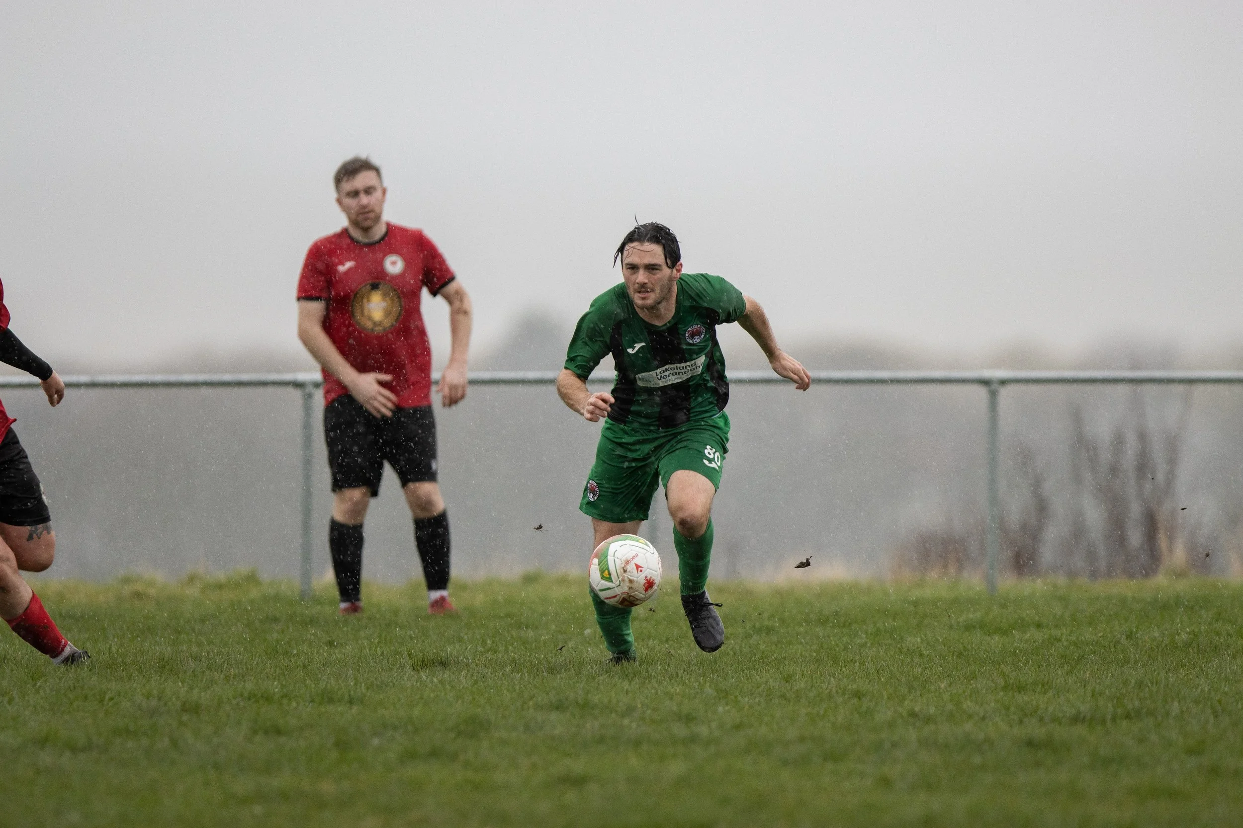 A soccer player in a green uniform running with a soccer ball on a grassy field in rainy weather, two players in red uniforms and black shorts are in the background.