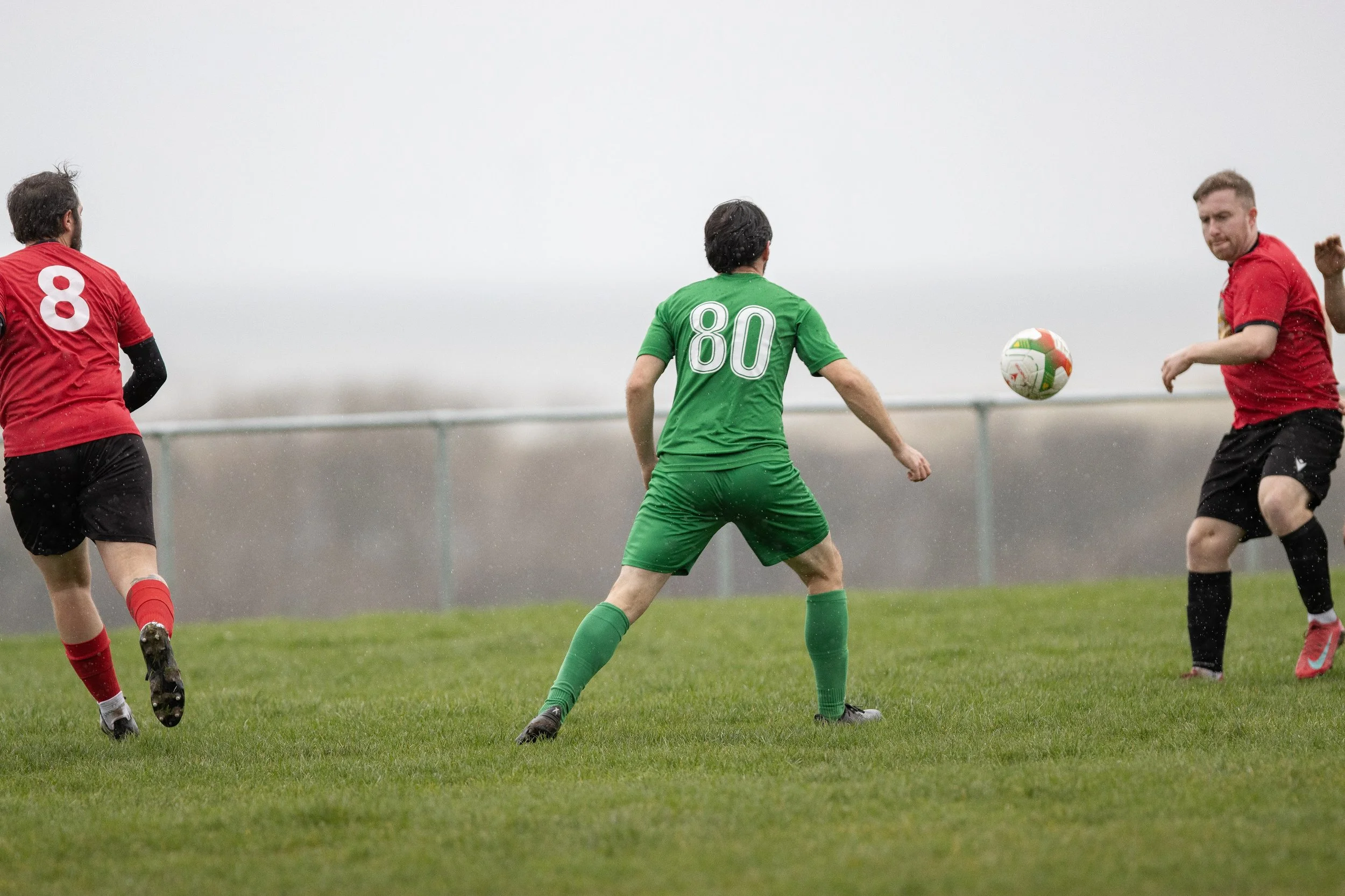 Soccer match on a rainy day with players in red and green jerseys, focusing on players near the ball.