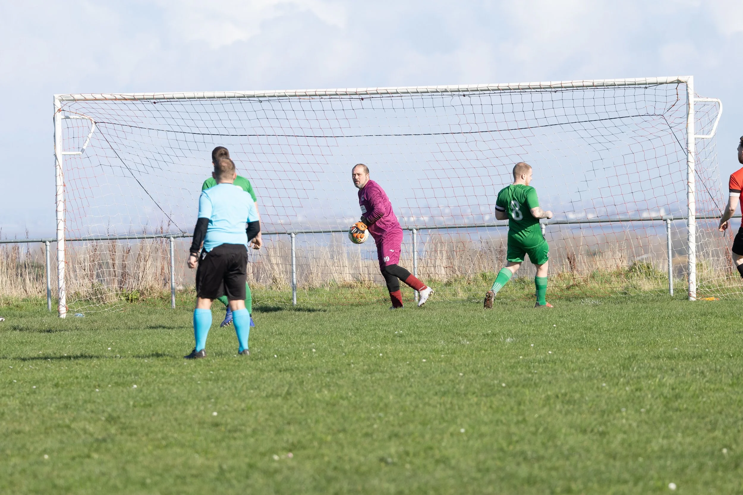 Soccer players on the field near the goal, with a goalkeeper in purple holding a soccer ball, and other players nearby, during a match on a grassy field under a partly cloudy sky.