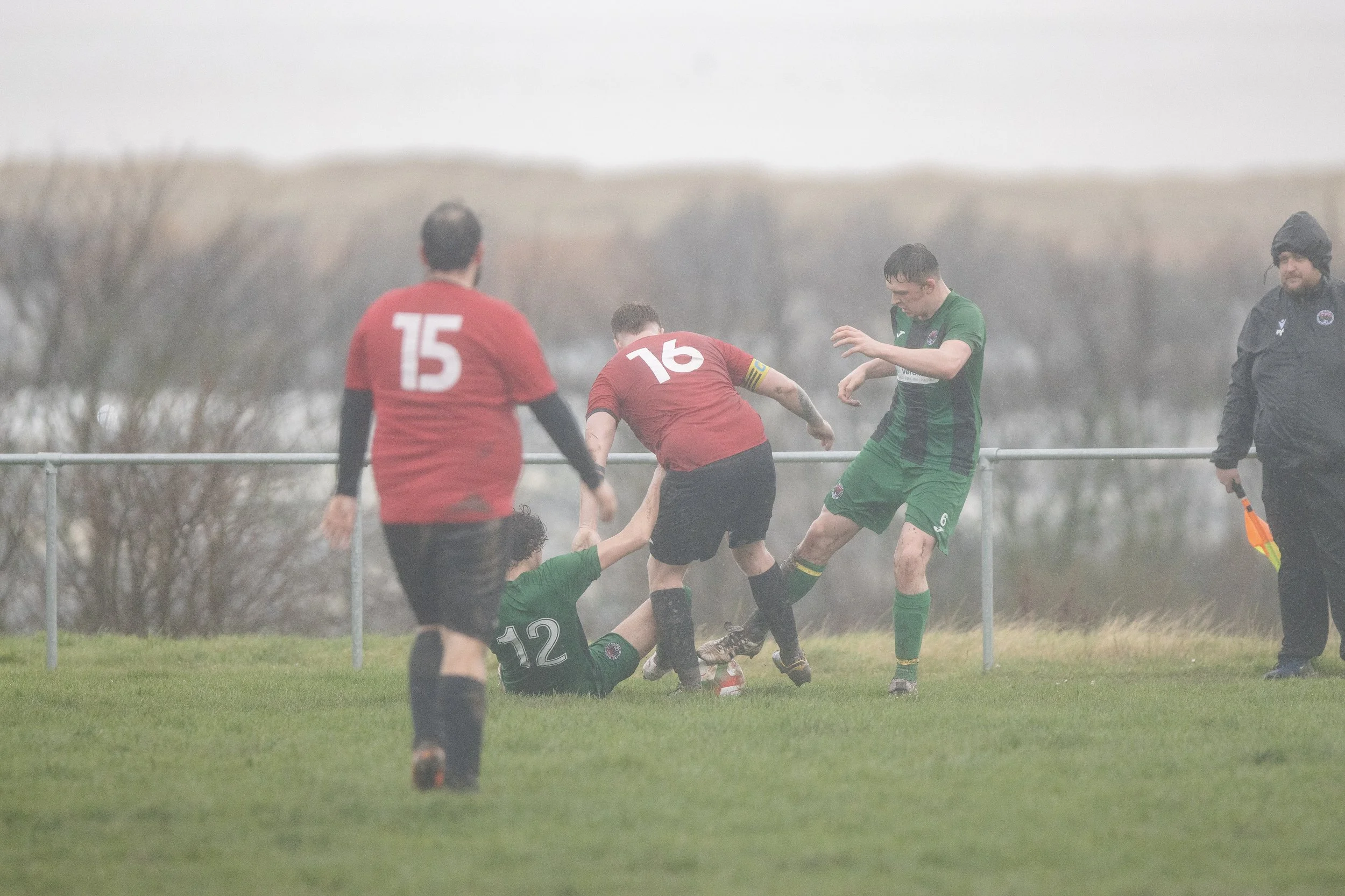 Rugby players in green and red jerseys competing for the ball during a match in rainy weather, with an official standing nearby.