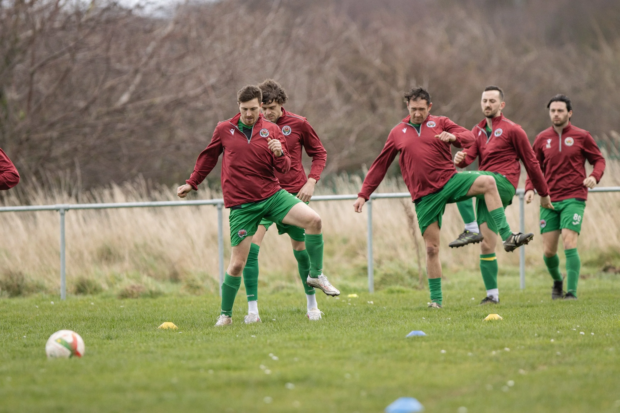 Soccer players in maroon and green uniforms practicing on a grassy field with cones, with a fence and trees in the background.