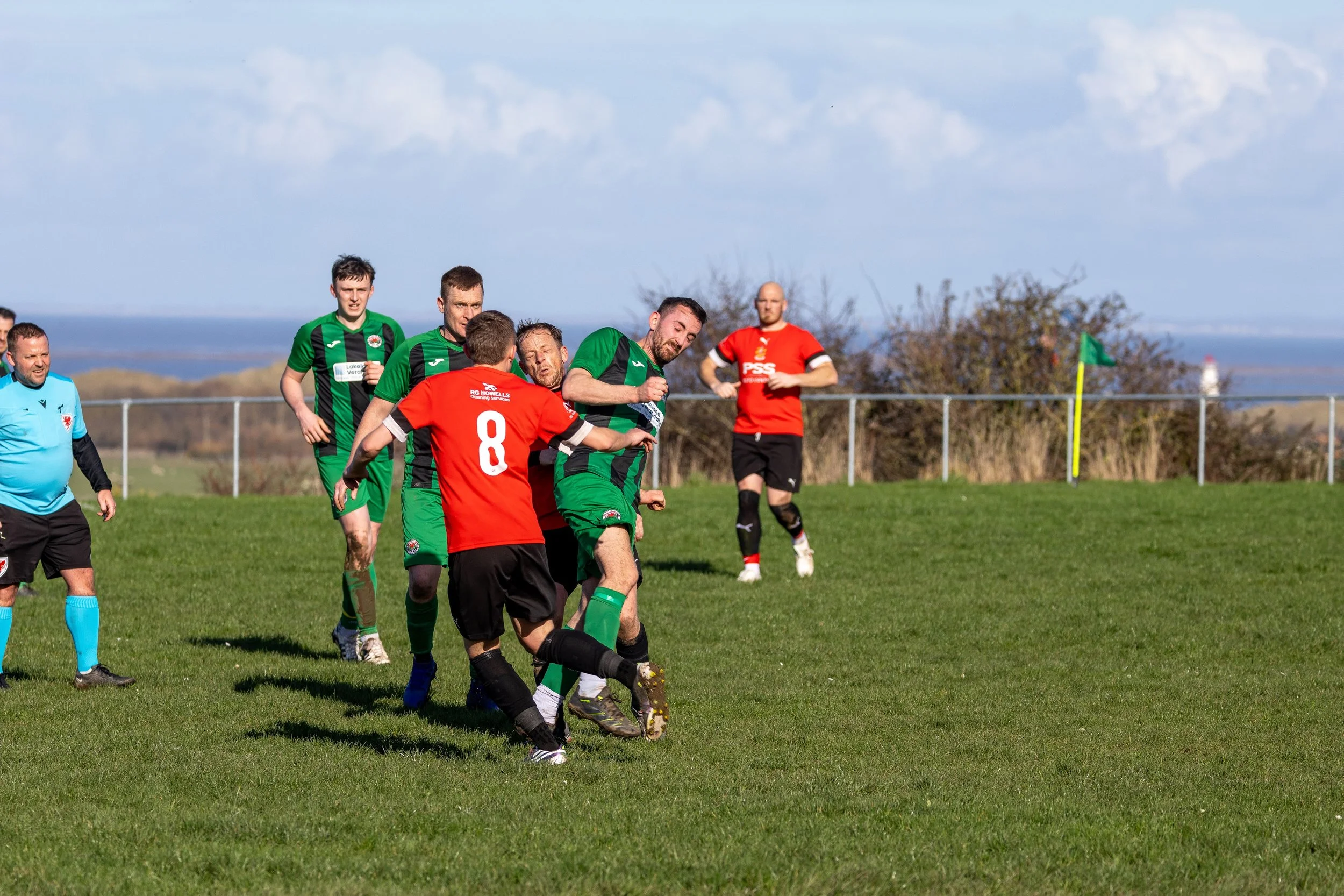 Soccer players in green and red jerseys compete for the ball on a grassy field during a match, with a blue sky and distant landscape in the background.