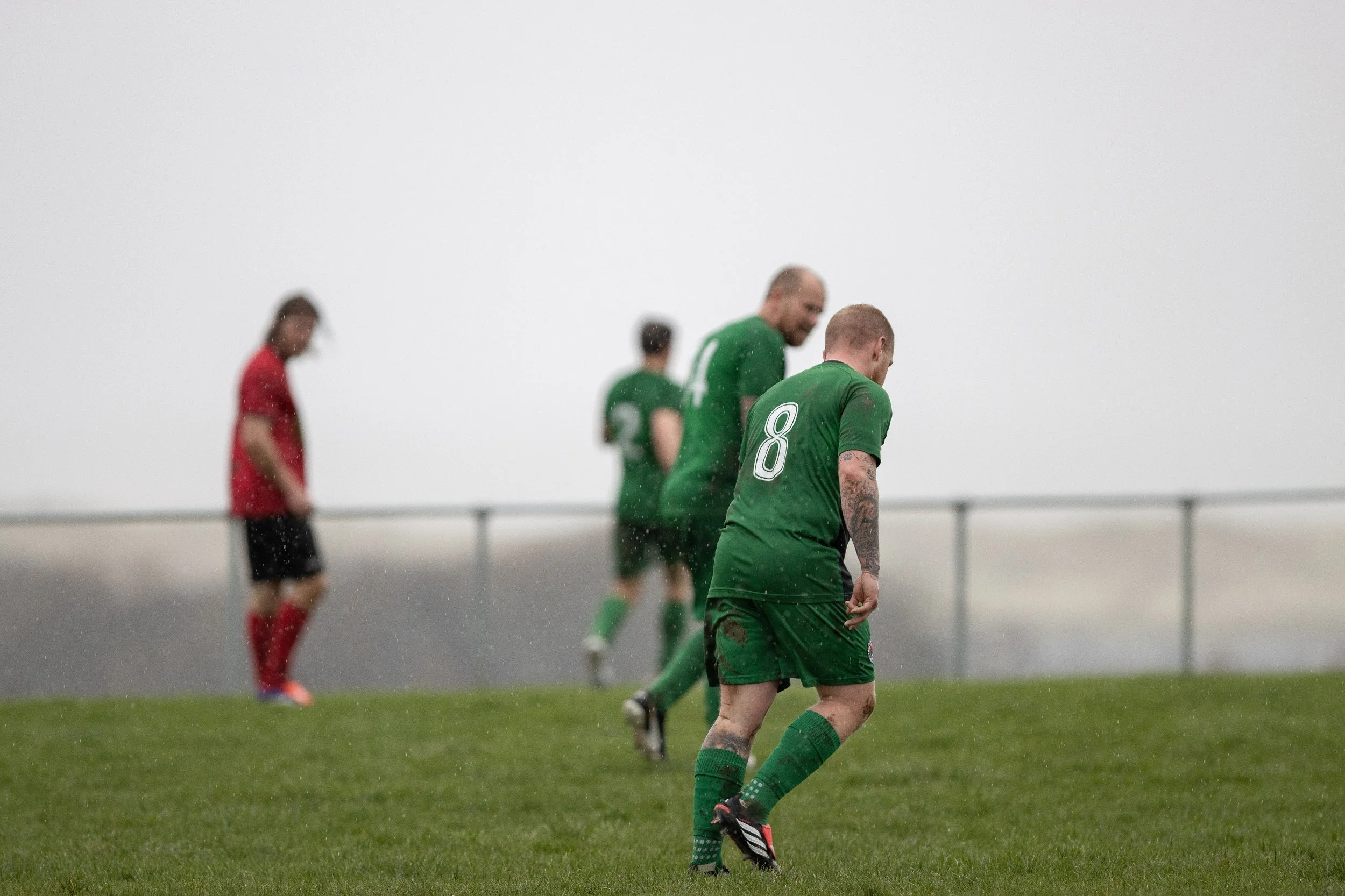 A soccer match on a rainy day with four players on the field, three in green jerseys and one in a red jersey, on a grassy field with a foggy background.