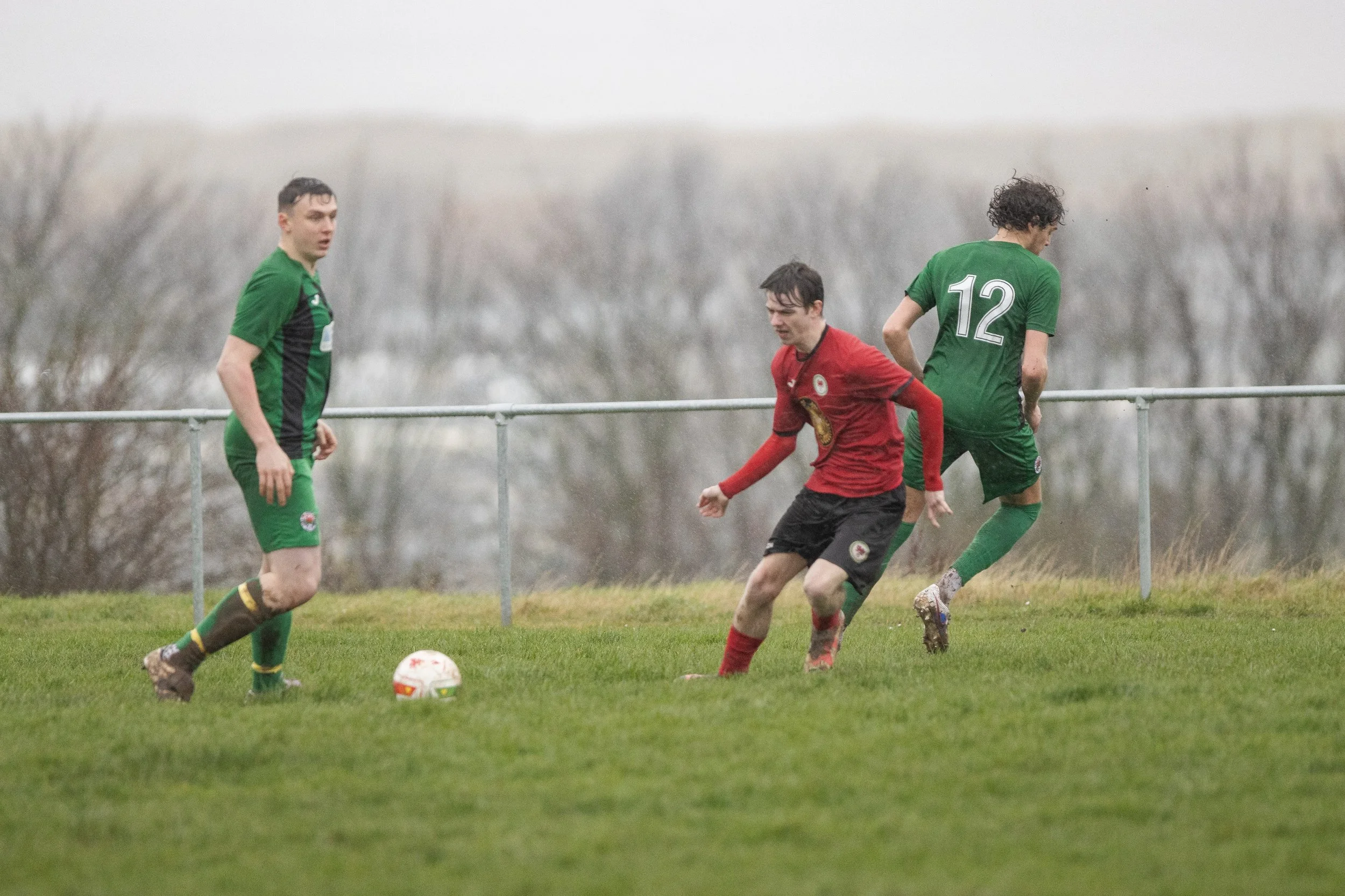 Three soccer players on a grassy field during a match, with one in a red uniform kicking the ball and two in green uniforms nearby.
