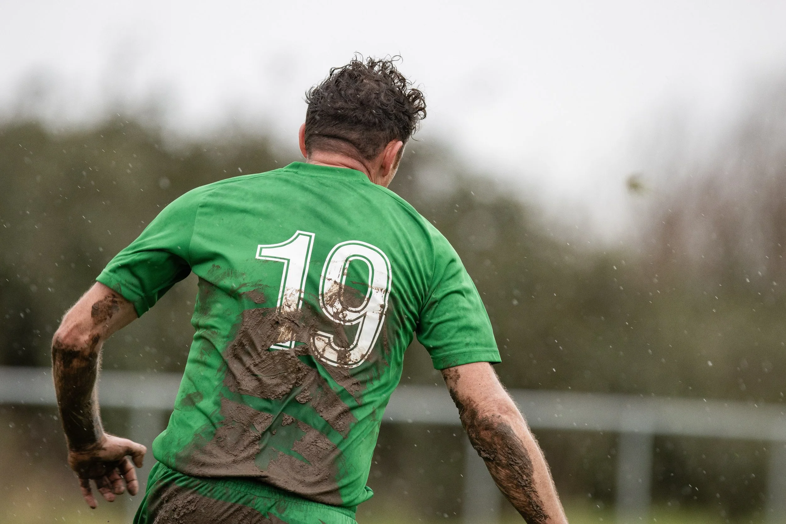 A soccer player wearing a green jersey with the number 19, covered in mud, during a match in rainy weather.