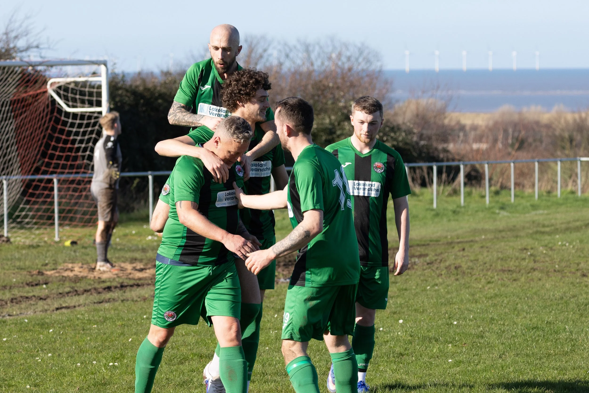 Soccer players in green jerseys celebrating on a grass field with ocean and wind turbines in the background.