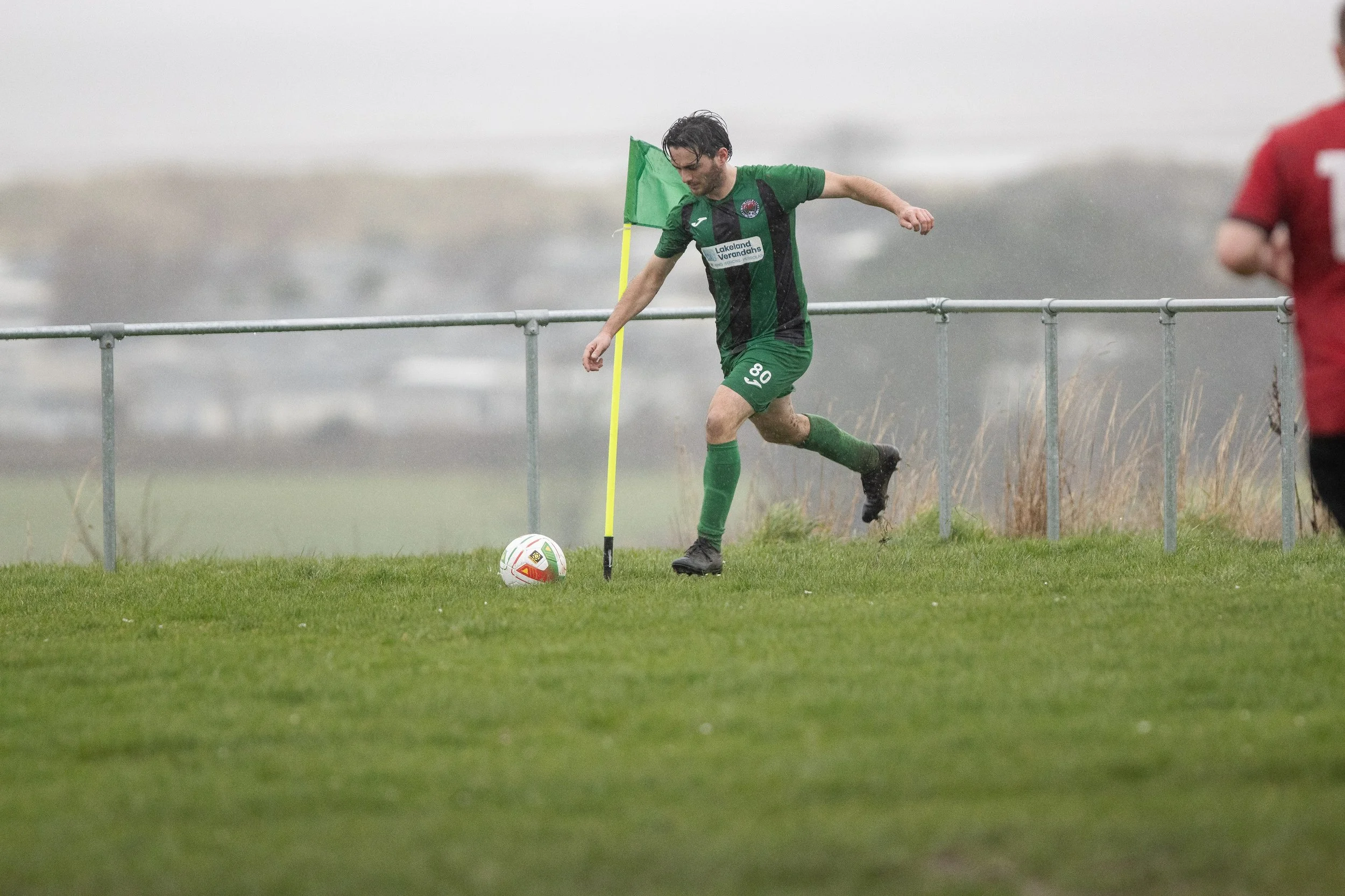 A soccer player dressed in a green uniform with the number 08 kicks a soccer ball on a grassy field near a metal railing, with a football flag in the background during rainy weather.