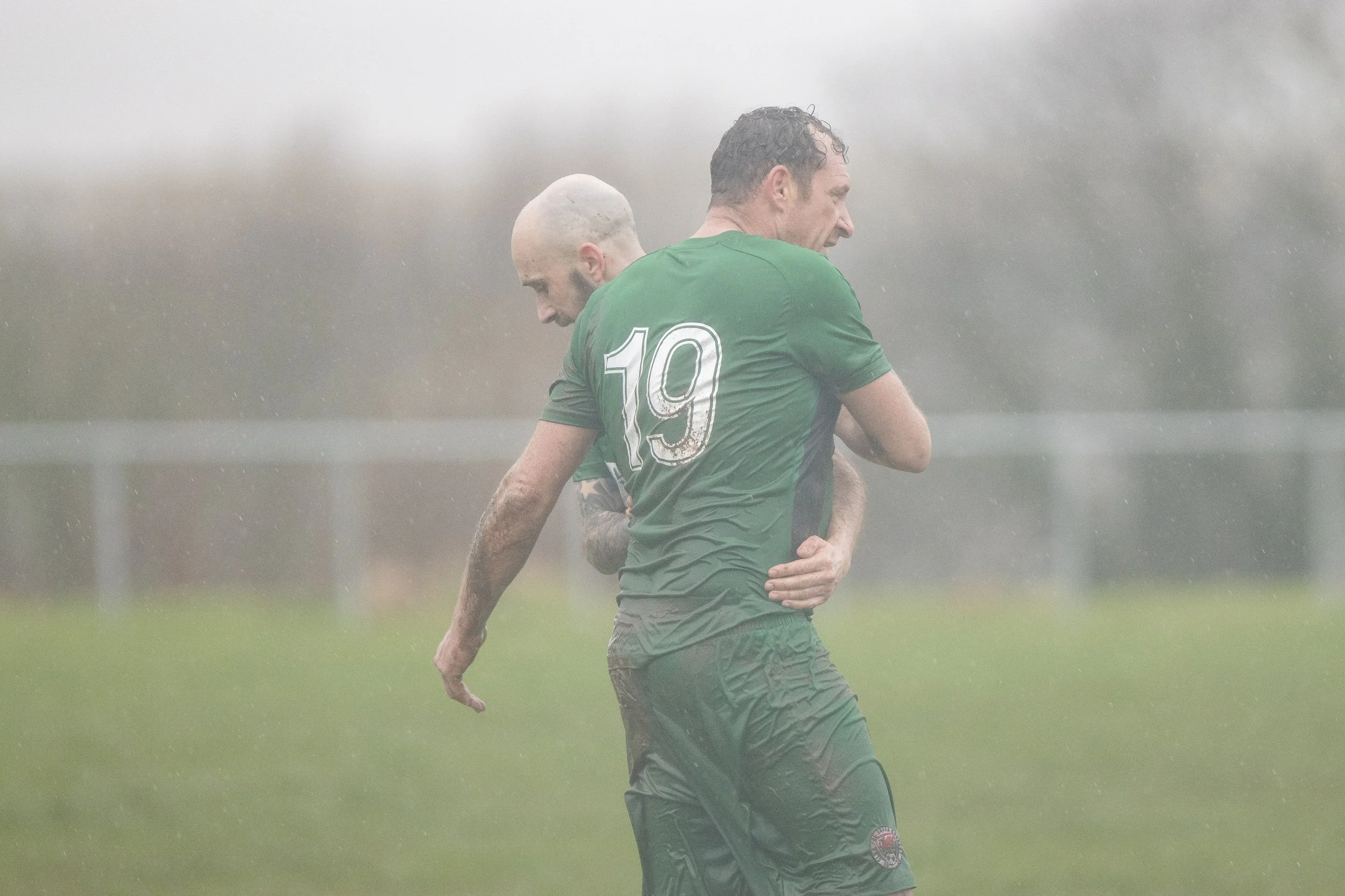 Two soccer players wearing green uniforms on a rain-soaked field, one with the number 19 on his back, celebrating after a match or goal.