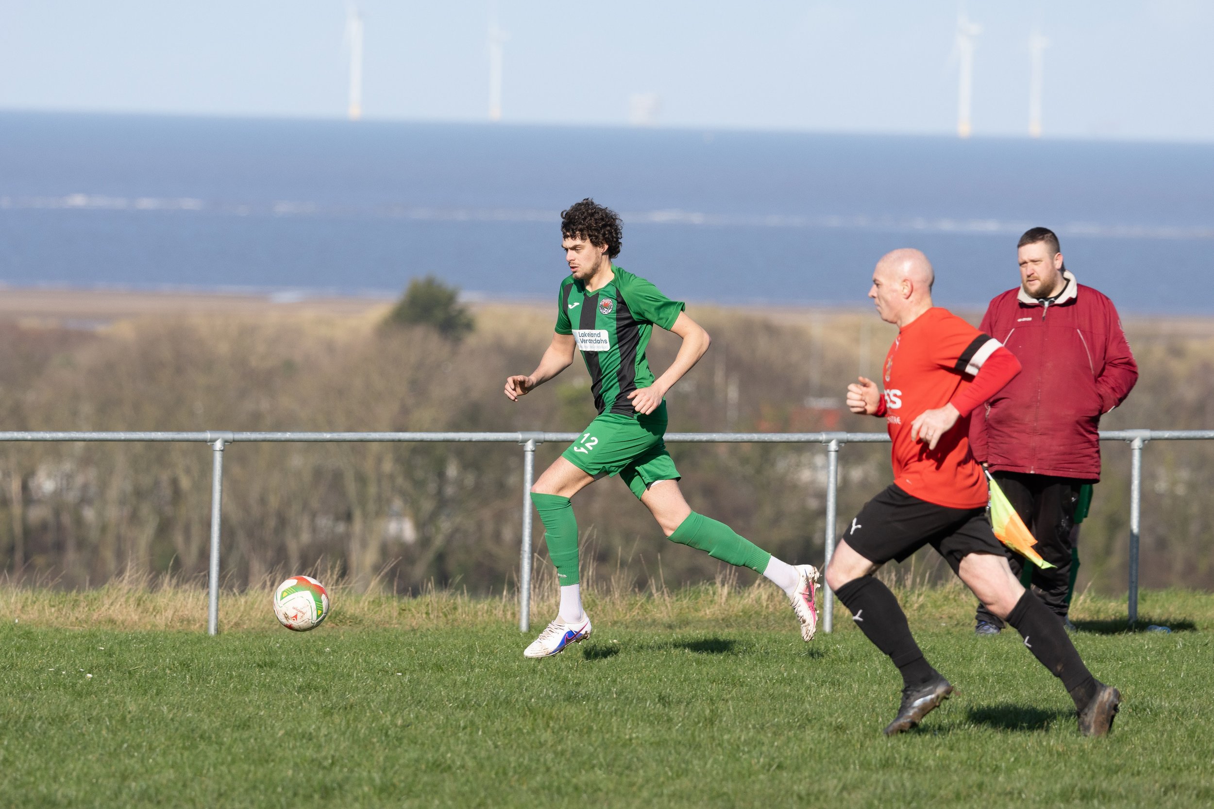 A soccer player in a green uniform is running after a ball on a grassy field, with two men in red and maroon jackets walking in the background against a backdrop of a body of water and a clear sky.