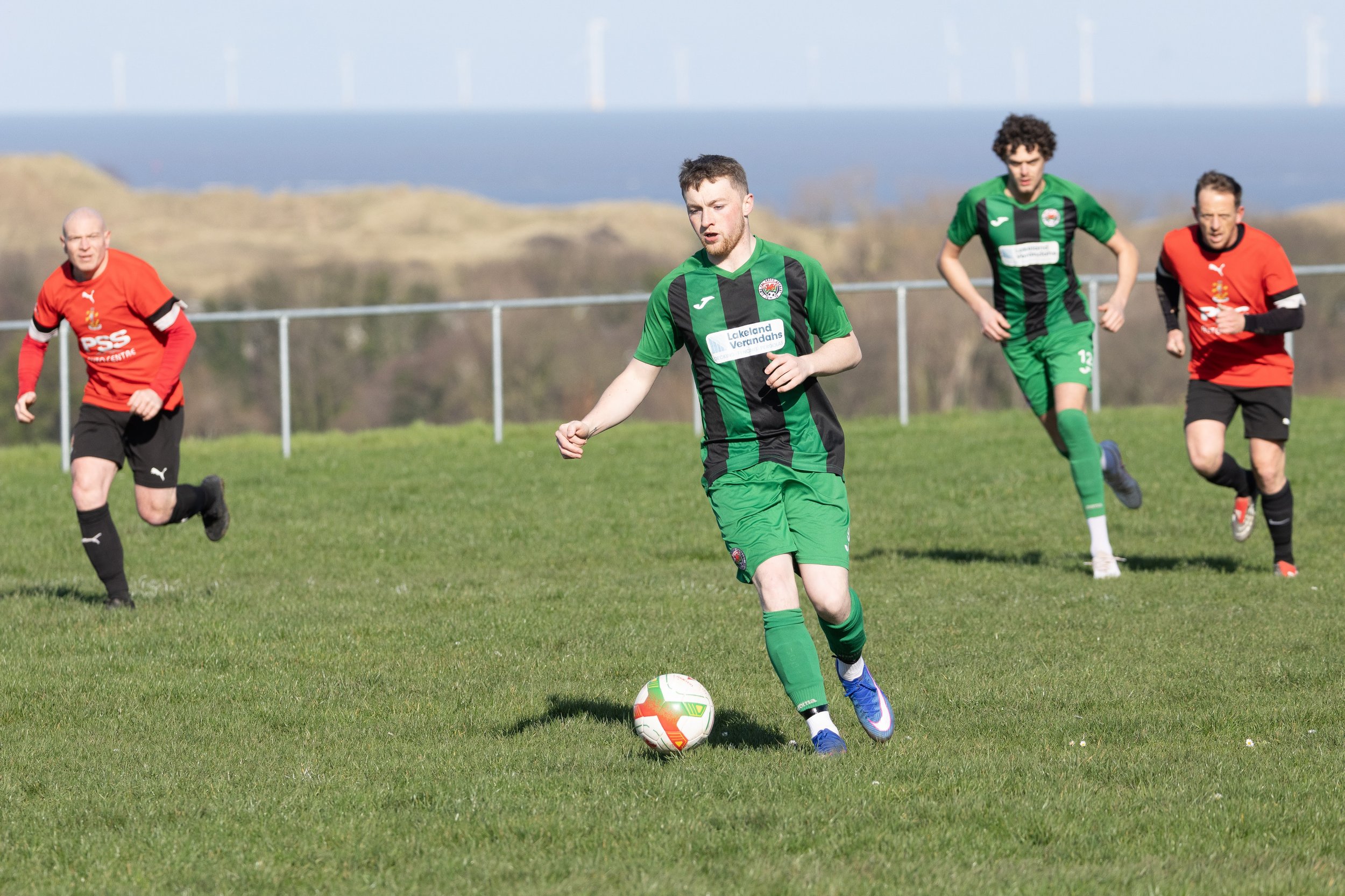 Soccer players on a grassy field during a match, with one player in green controlling the ball, and two opponents in red running behind.