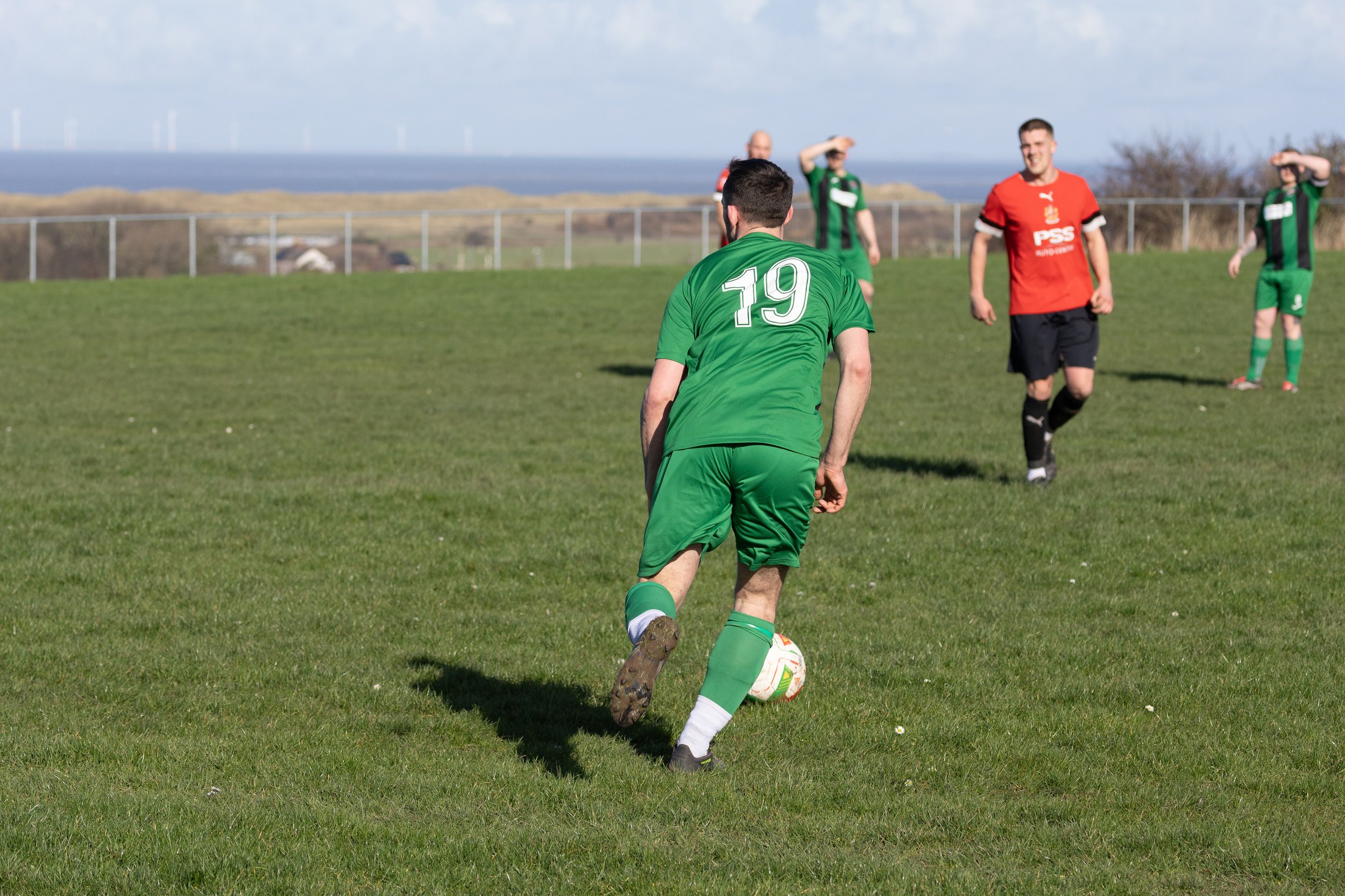 Soccer player in a green uniform with the number 19 about to kick a soccer ball on a grassy field, with other players in the background and a scenic landscape with wind turbines.