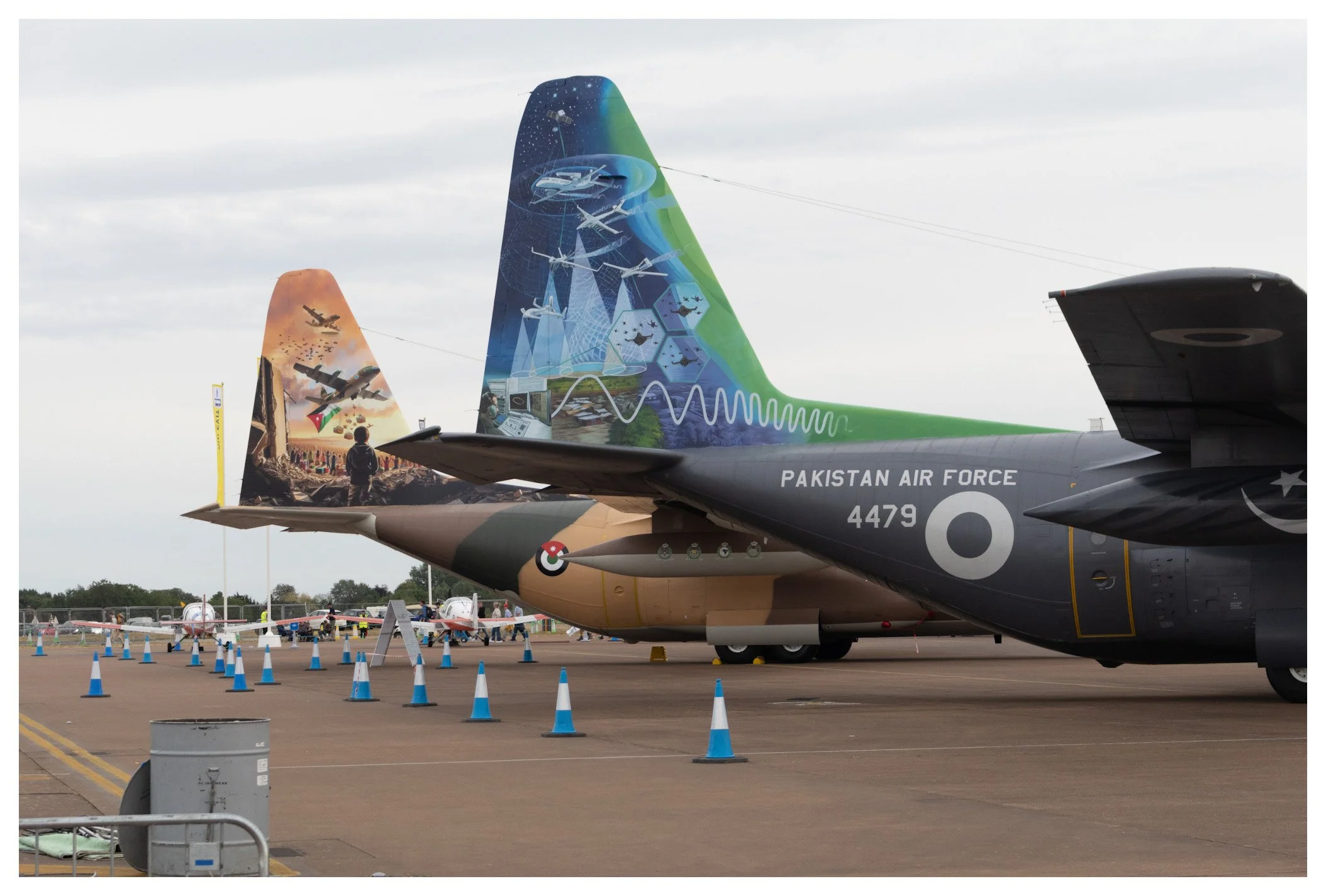Two Pakistan Air Force fighter jets parked on an airfield, decorated with colorful murals on their tails, featuring military themes and technical illustrations, surrounded by orange and blue cones.