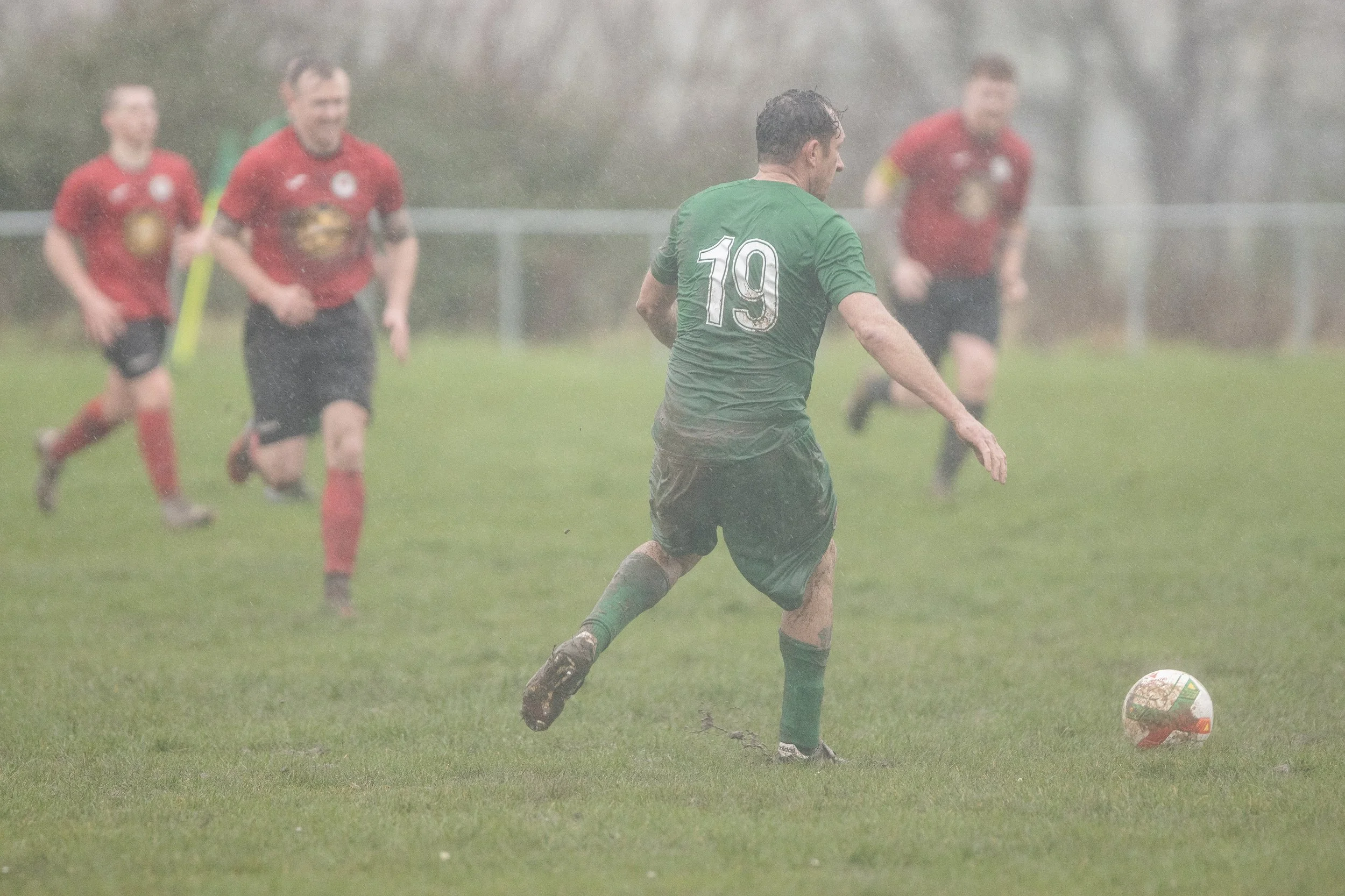 A soccer player wearing a green jersey with the number 19 is running in the rain, chasing a soccer ball, while three players in red jerseys are running behind him on a grassy field.