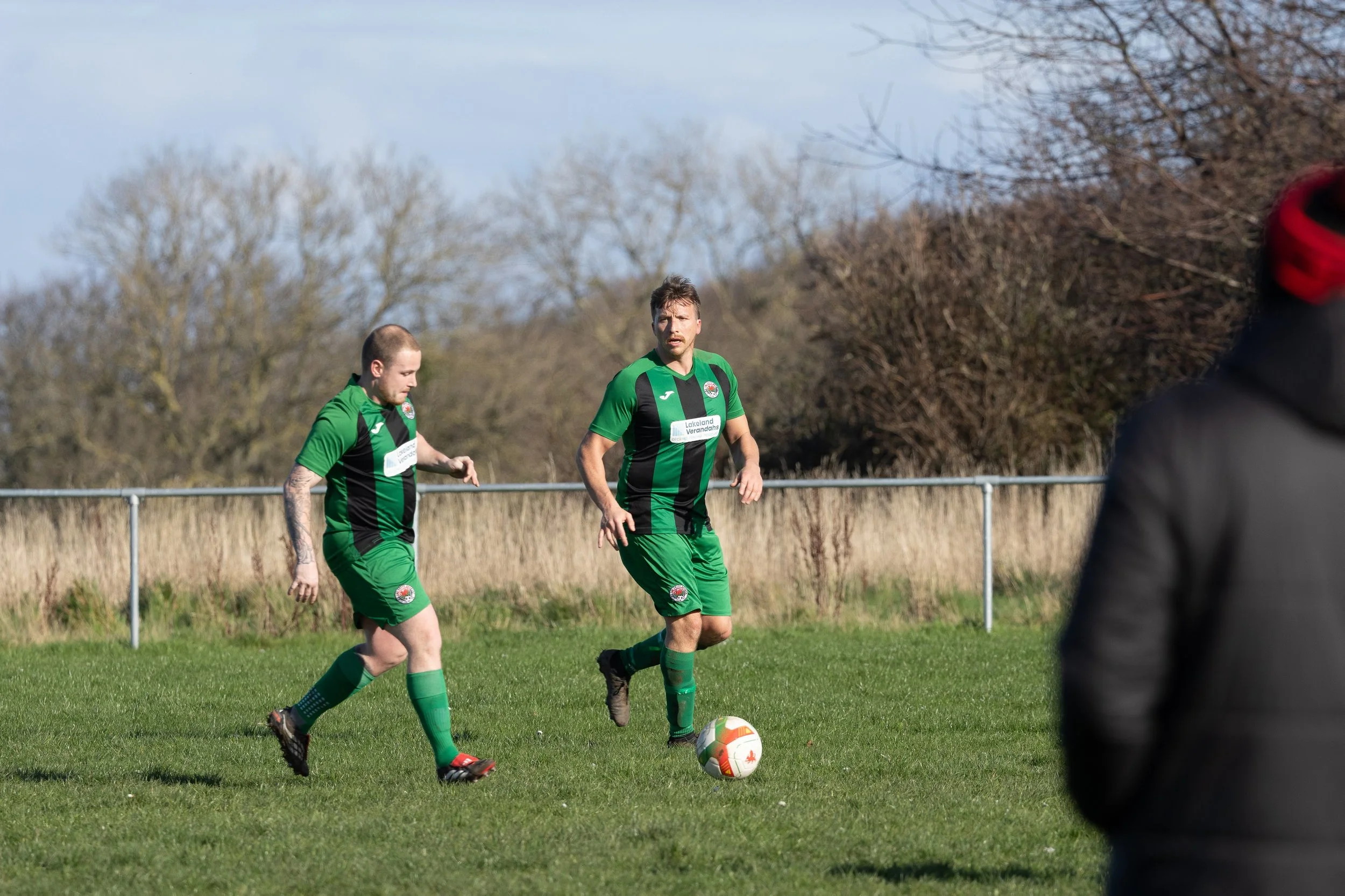 Two soccer players in green and black uniforms on a grassy field during a game, one with the ball, with a person in a black jacket observing on the right side.