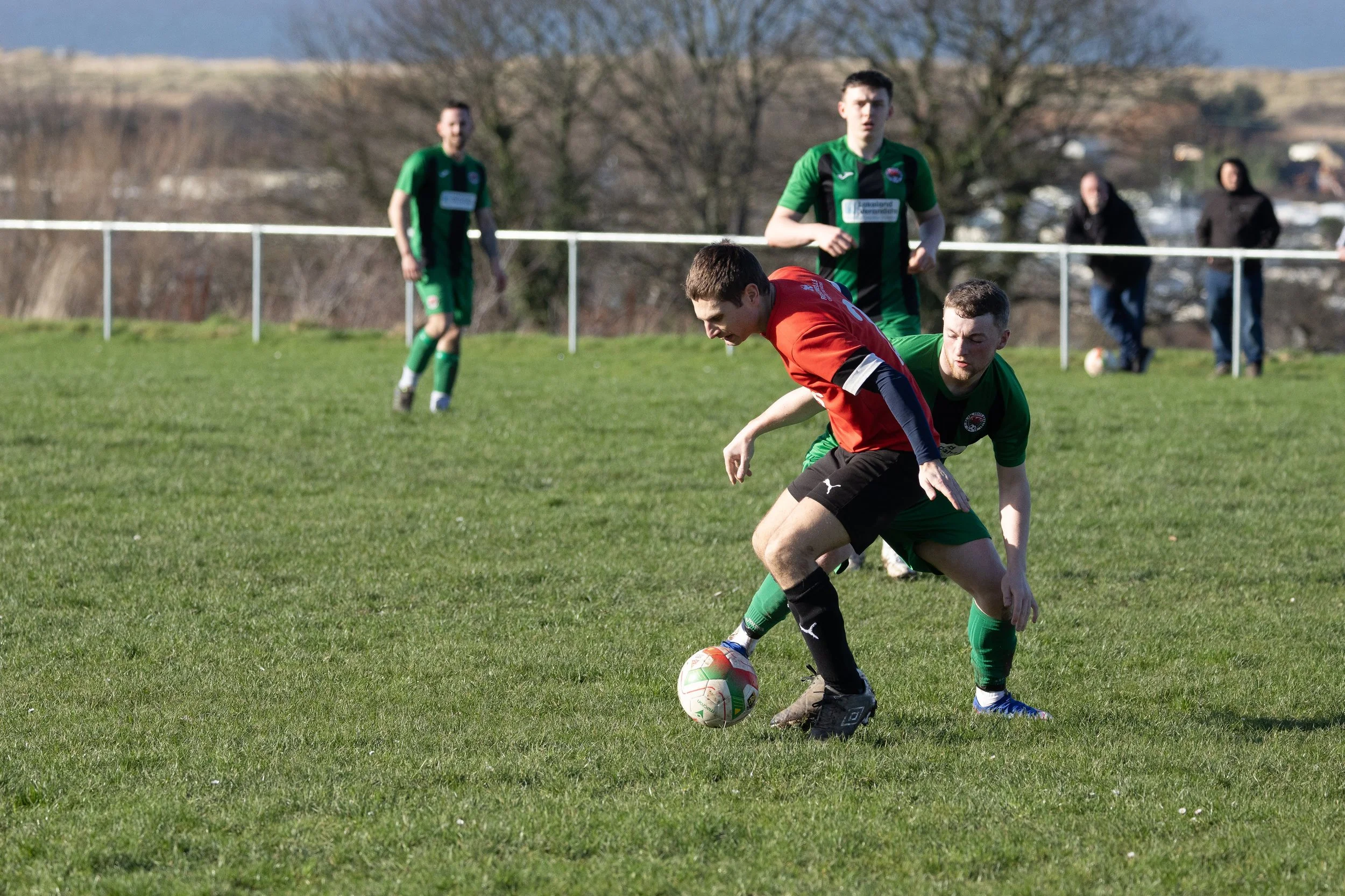 Two soccer players in green uniforms and one in a red and black uniform fight for the ball on a grassy field, with two spectators and trees in the background.