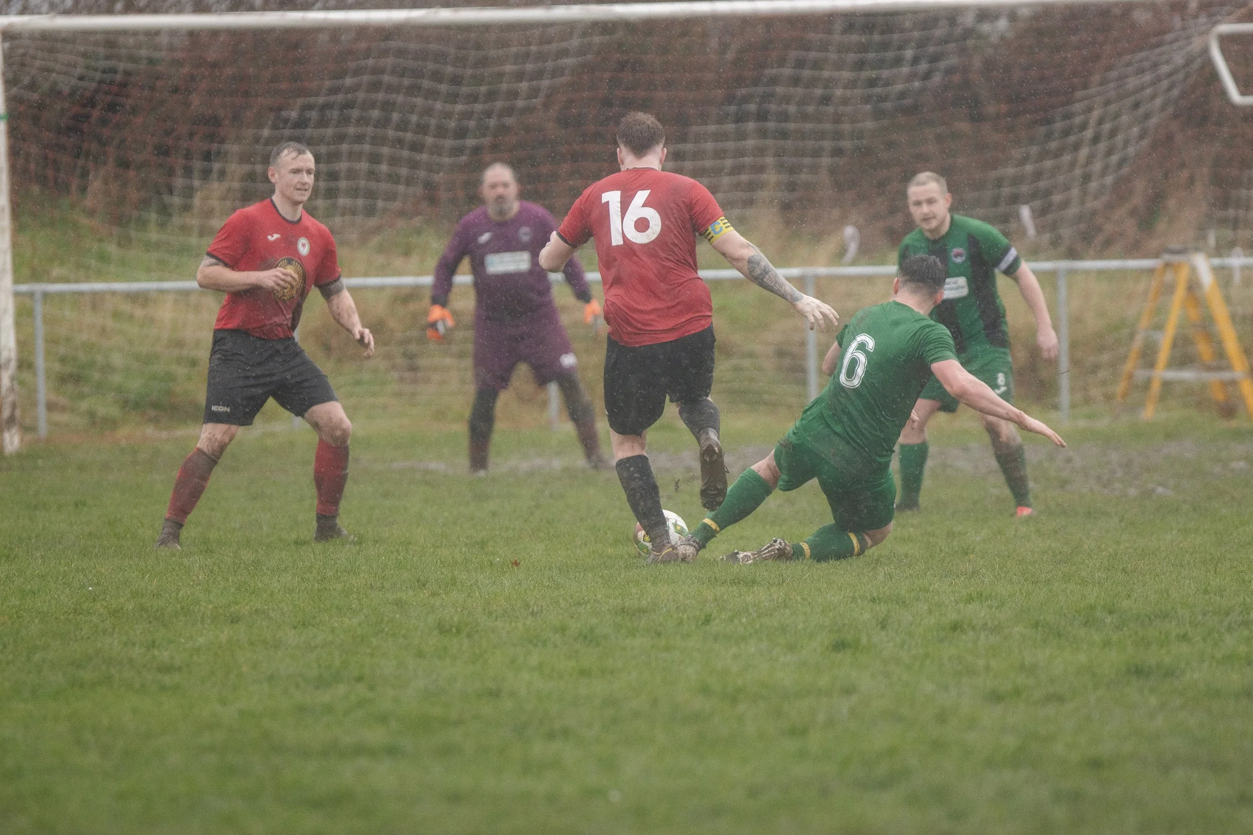 A soccer match taking place in rainy weather as players chase the ball near the goal. One player in green is sliding to kick the ball while players in red are defending.