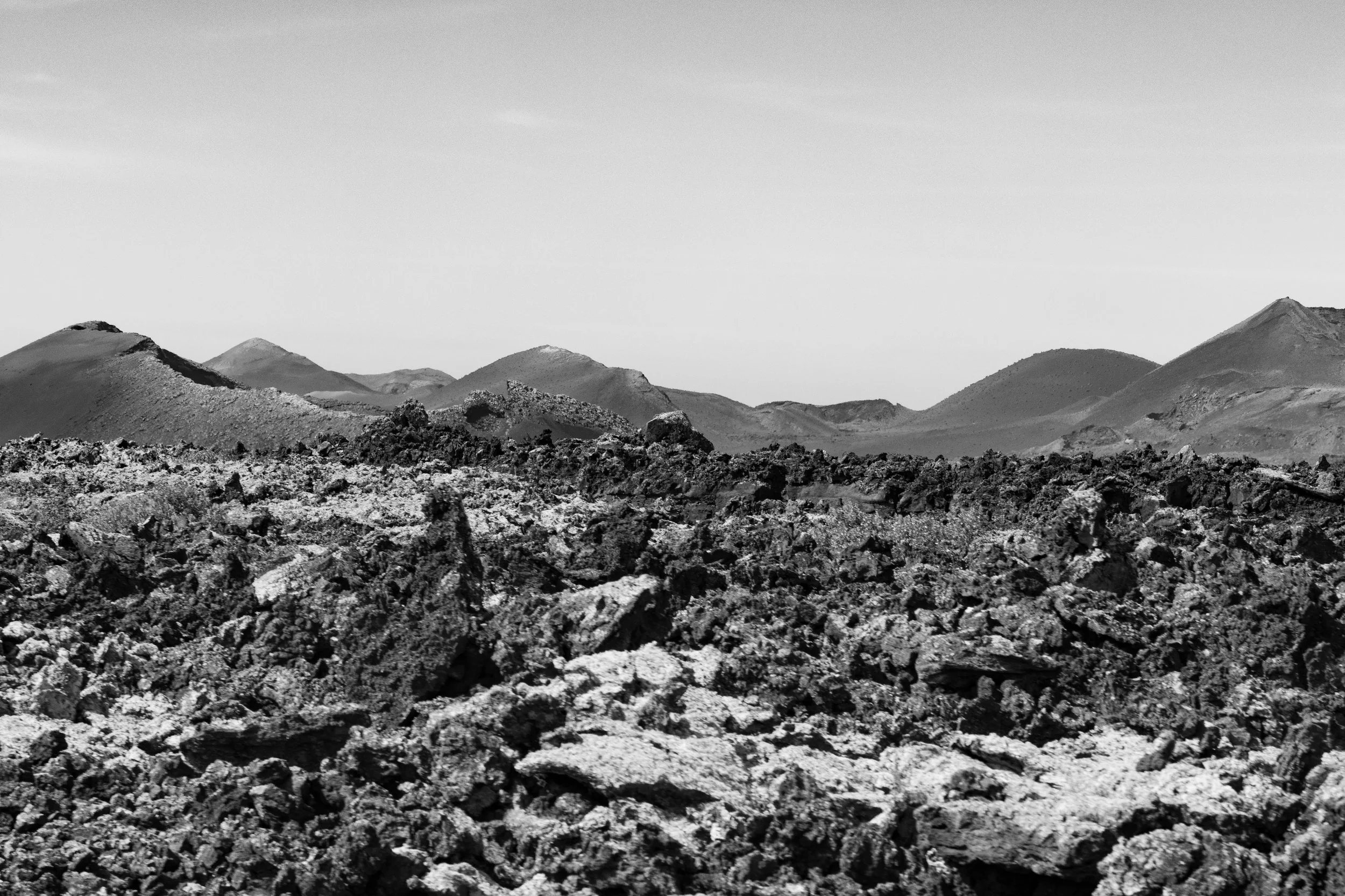 Black and white photo of a rugged volcanic landscape with rough, uneven rocks in the foreground and distant volcanic mountains under a clear sky.
