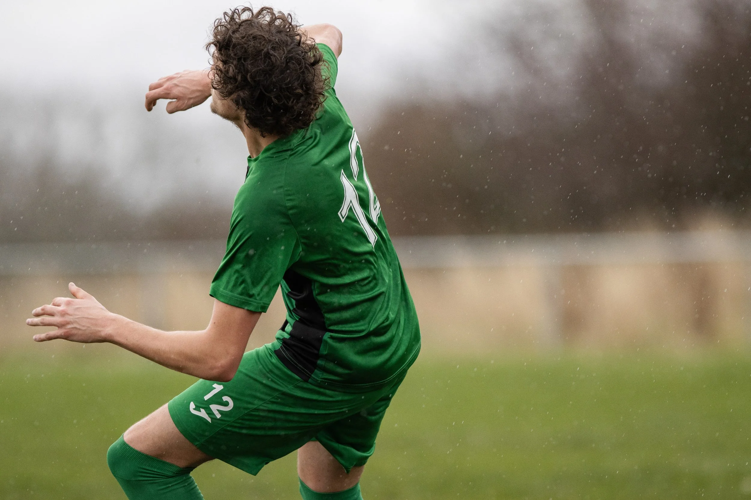 A soccer player in a green uniform with the number 12 is kicking a ball on a wet field during a rainy day.