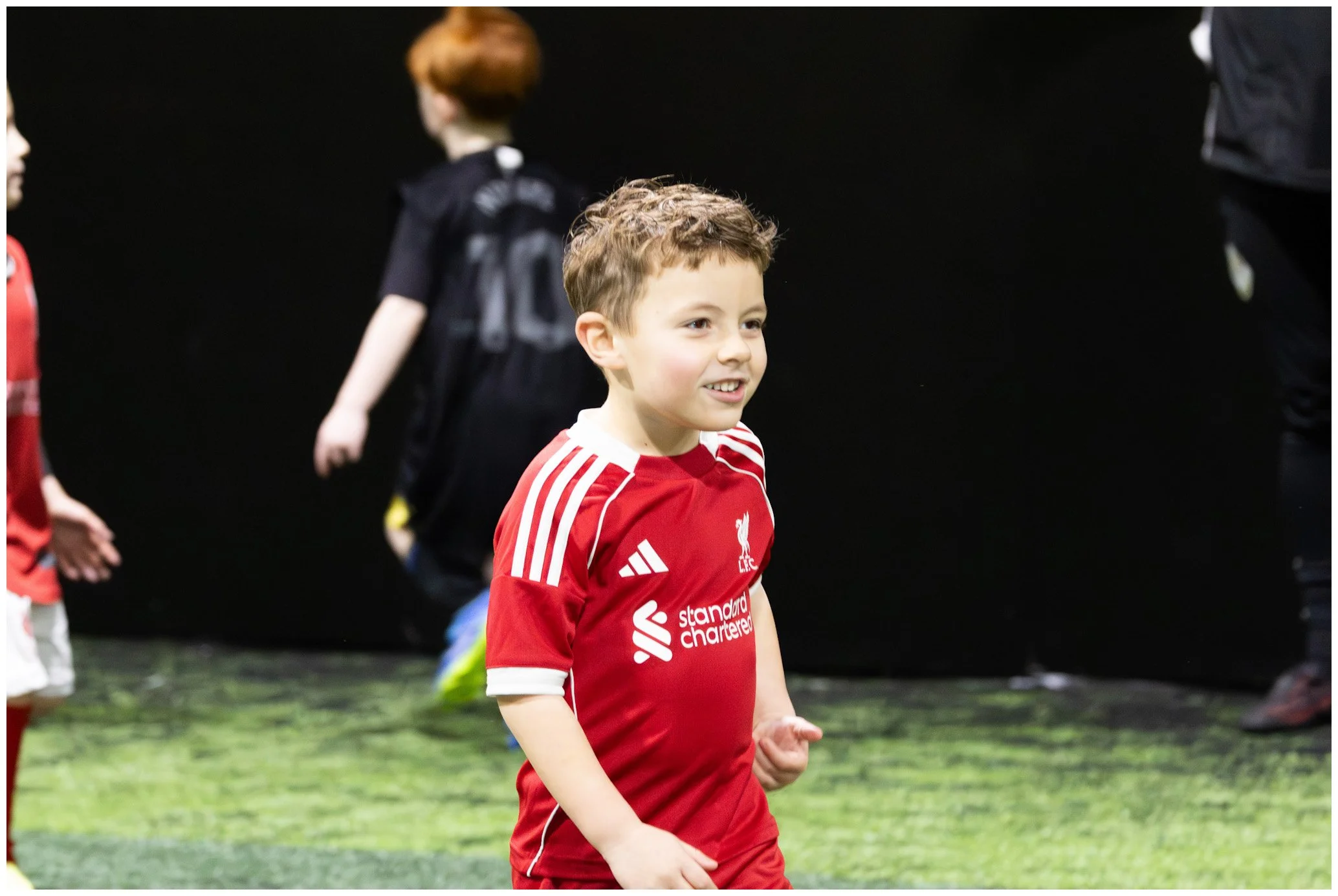 A young boy with curly brown hair smiling while wearing a red Liverpool football jersey, standing on a green indoor soccer field with other children and adults in the background.
