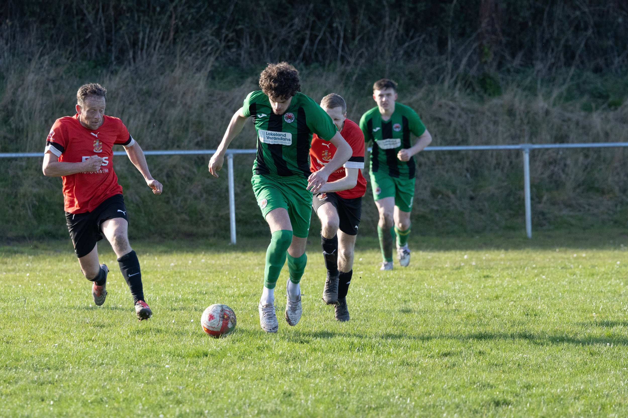 Four male soccer players running on a grassy field chasing the ball, with two players in red jerseys and two in green jerseys.
