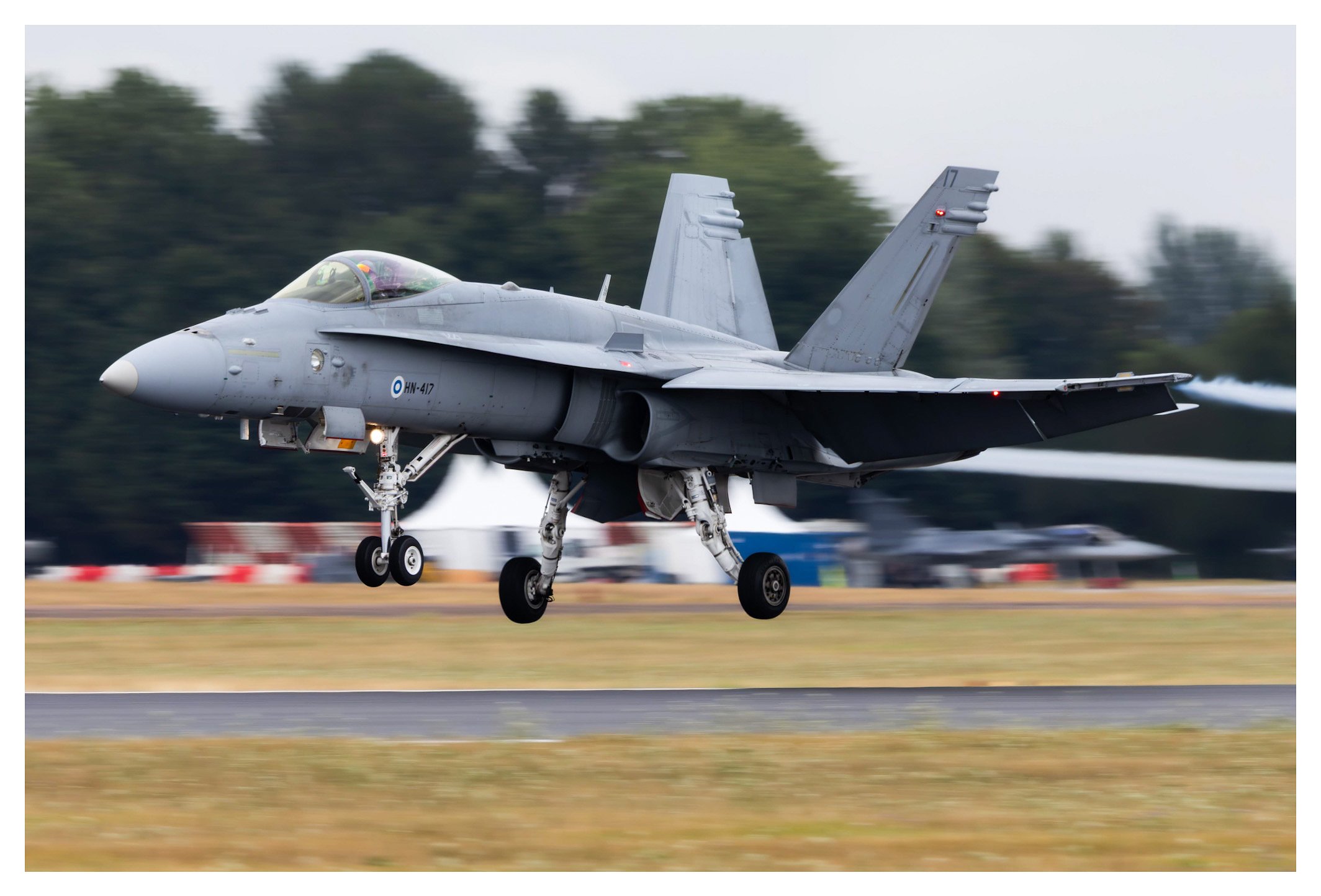 F-18 fighter jet flying low over runway with landing gear extended, kicking up dust and smoke, with blurred background of trees and people.